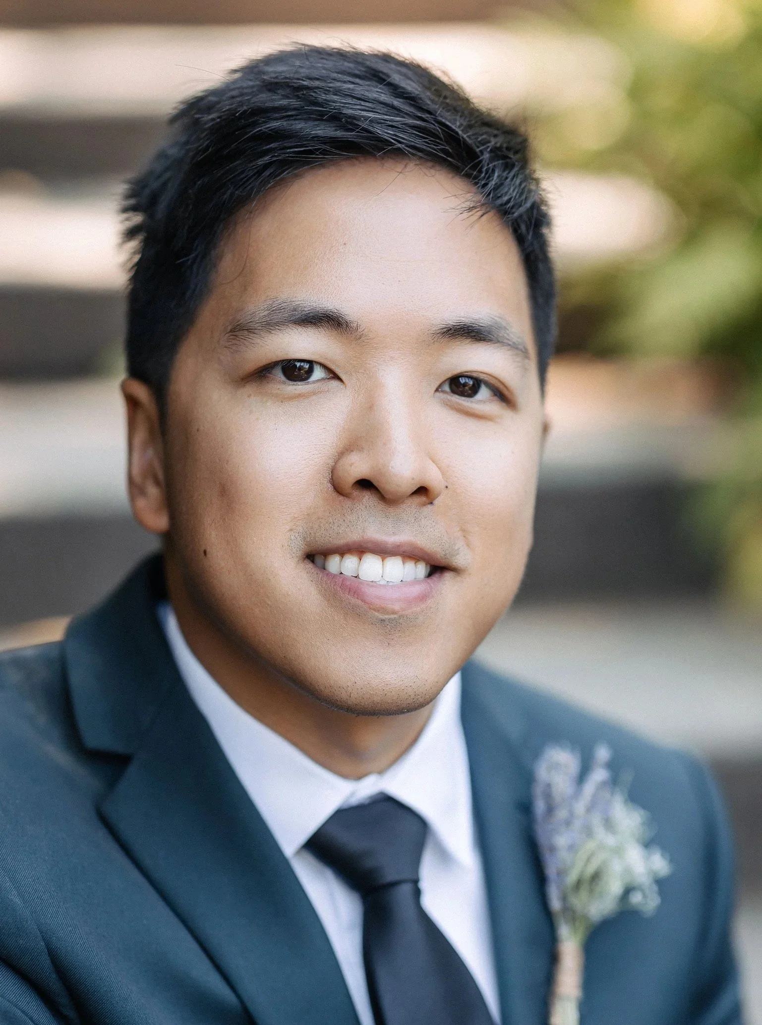 A young man in a navy blue suit with a white dress shirt and black tie, smiling outdoors with a blurred background of trees and greenery.