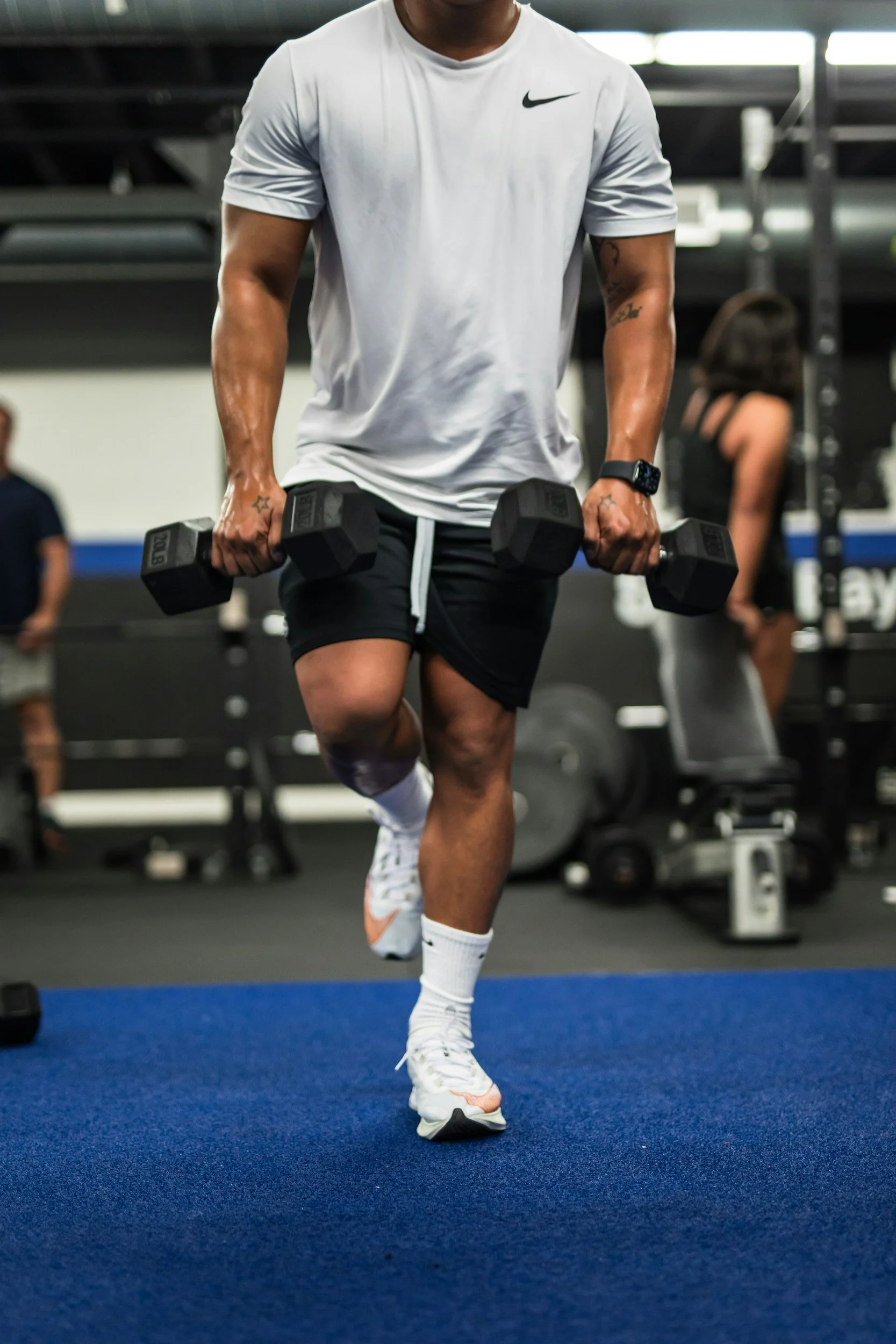 Person lifting dumbbells in a gym, wearing a white Nike t-shirt, black shorts, and white sports shoes.
