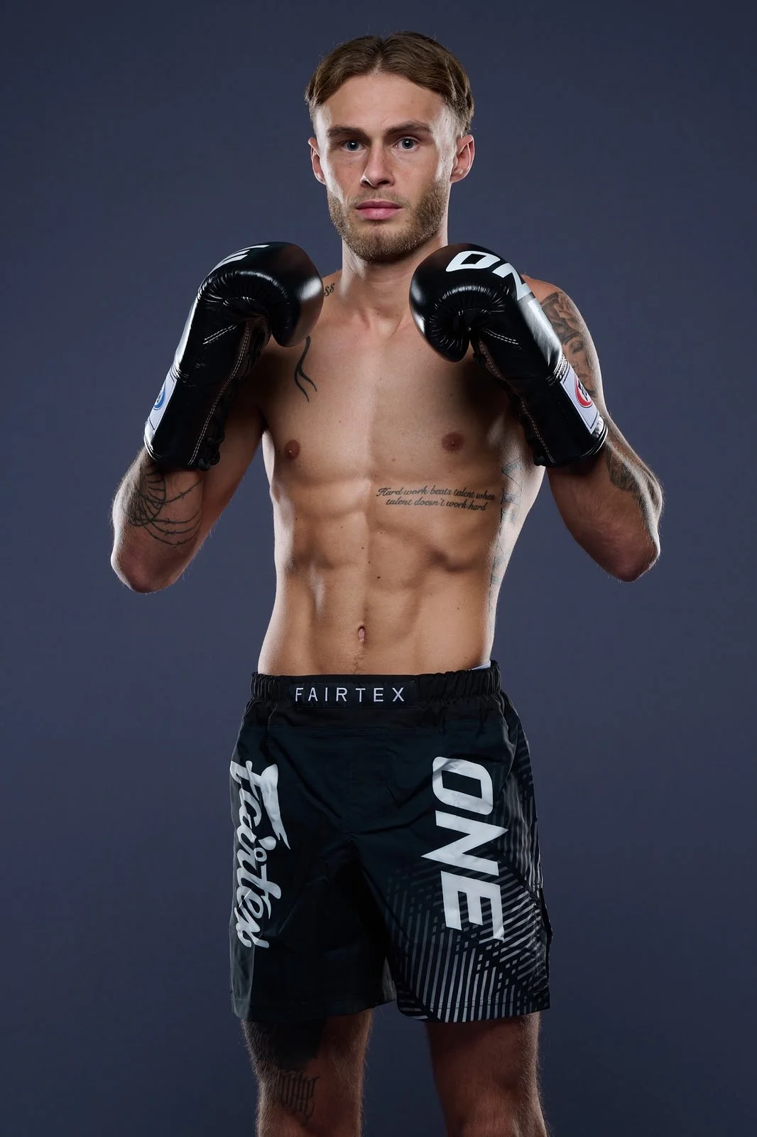 A young male boxer poses with fists raised, wearing black boxing gloves and shorts, shirtless, with tattoos visible on his arms and torso, against a plain dark gray background.