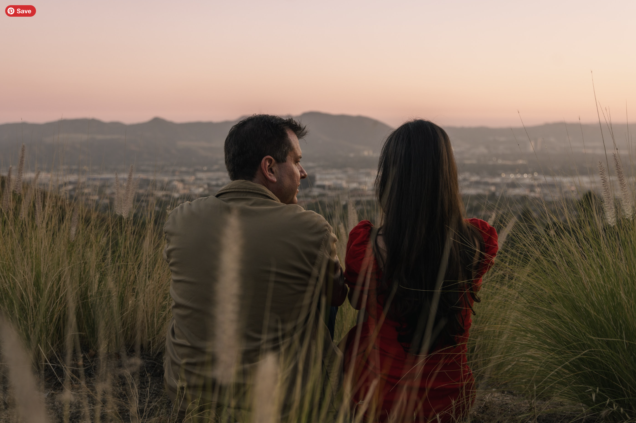 A man and a woman sitting amidst tall grasses, facing a distant view of mountains and a cityscape at sunset or sunrise.