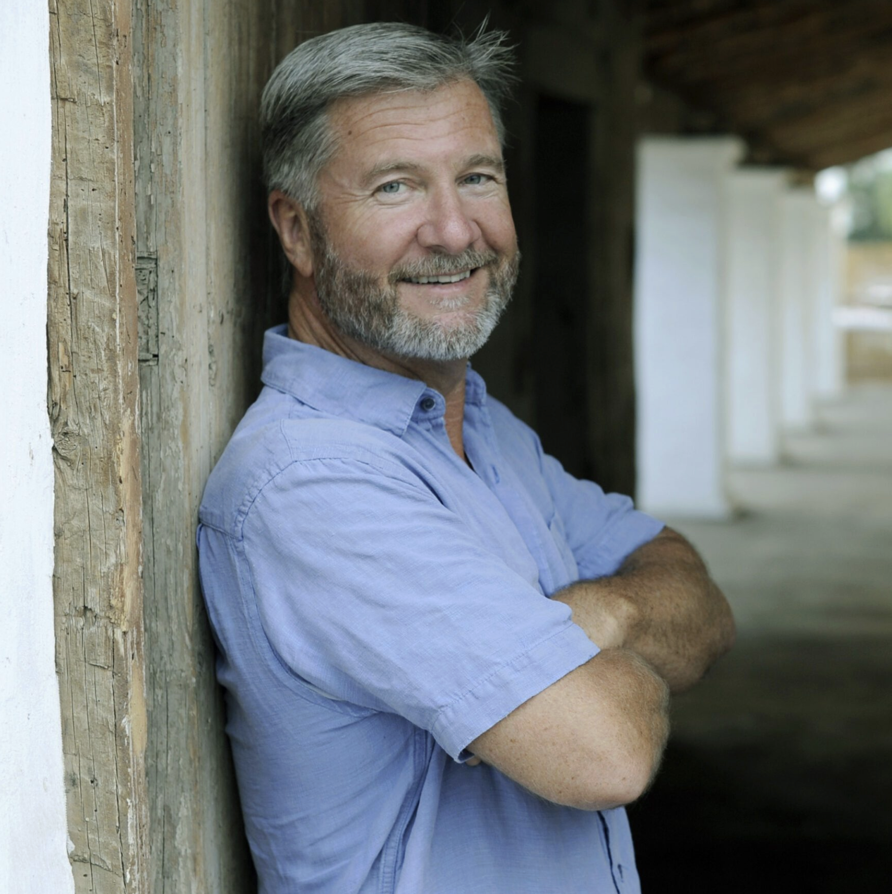 Professional headshot of Shelby Sim. A kind, grey-haired man in a blue polo, leaning against a slab of barn wood.