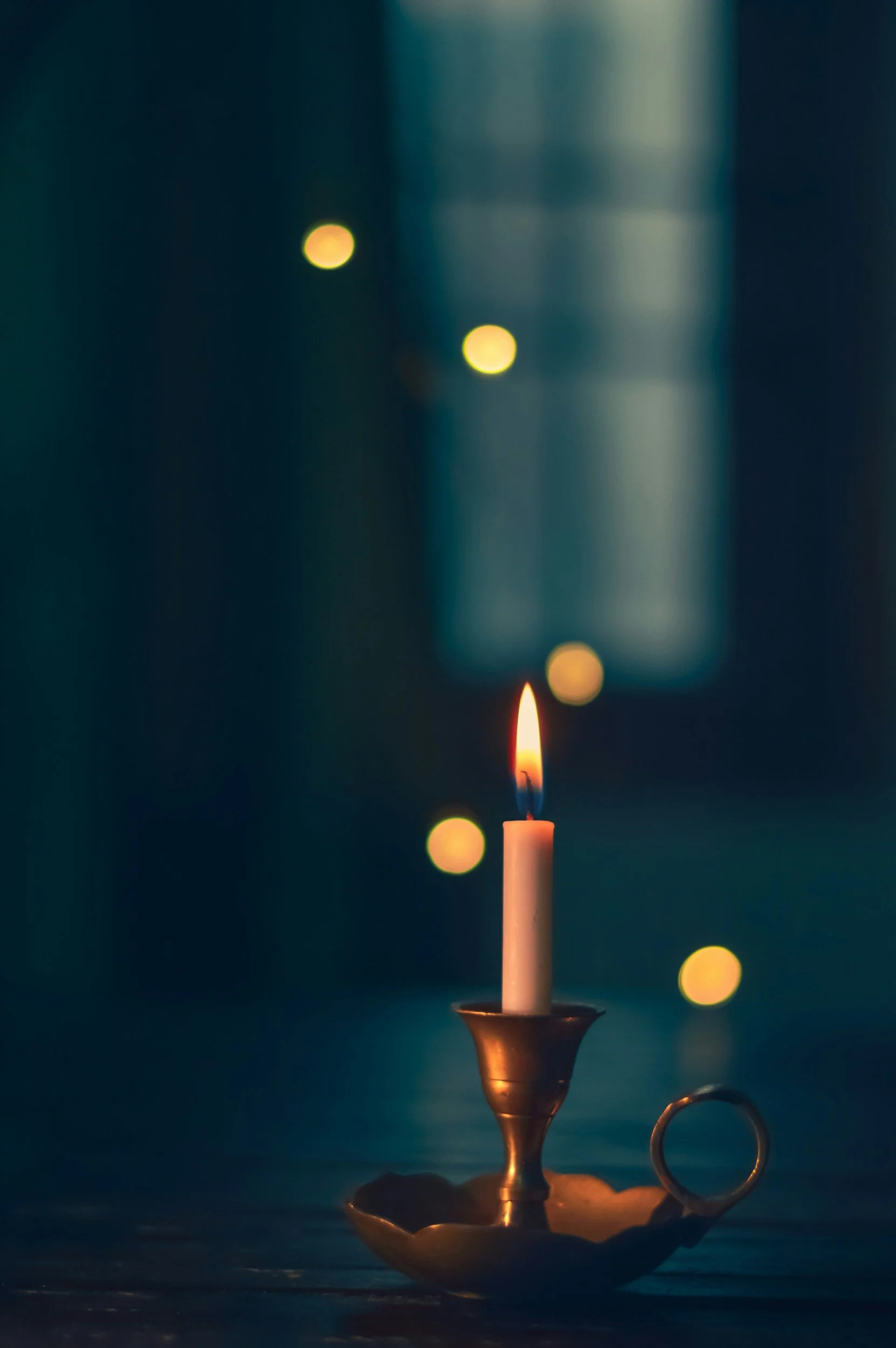 A lit white candle in a metal candle holder with a curved handle, placed on a dark wooden surface, with a dark teal background and blurred yellow lights.