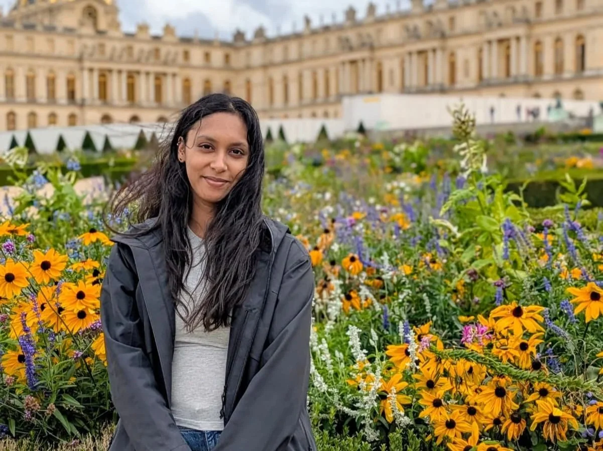 A woman with long black hair in a gray jacket and white shirt standing in a garden full of colorful flowers, with a historic building in the background.