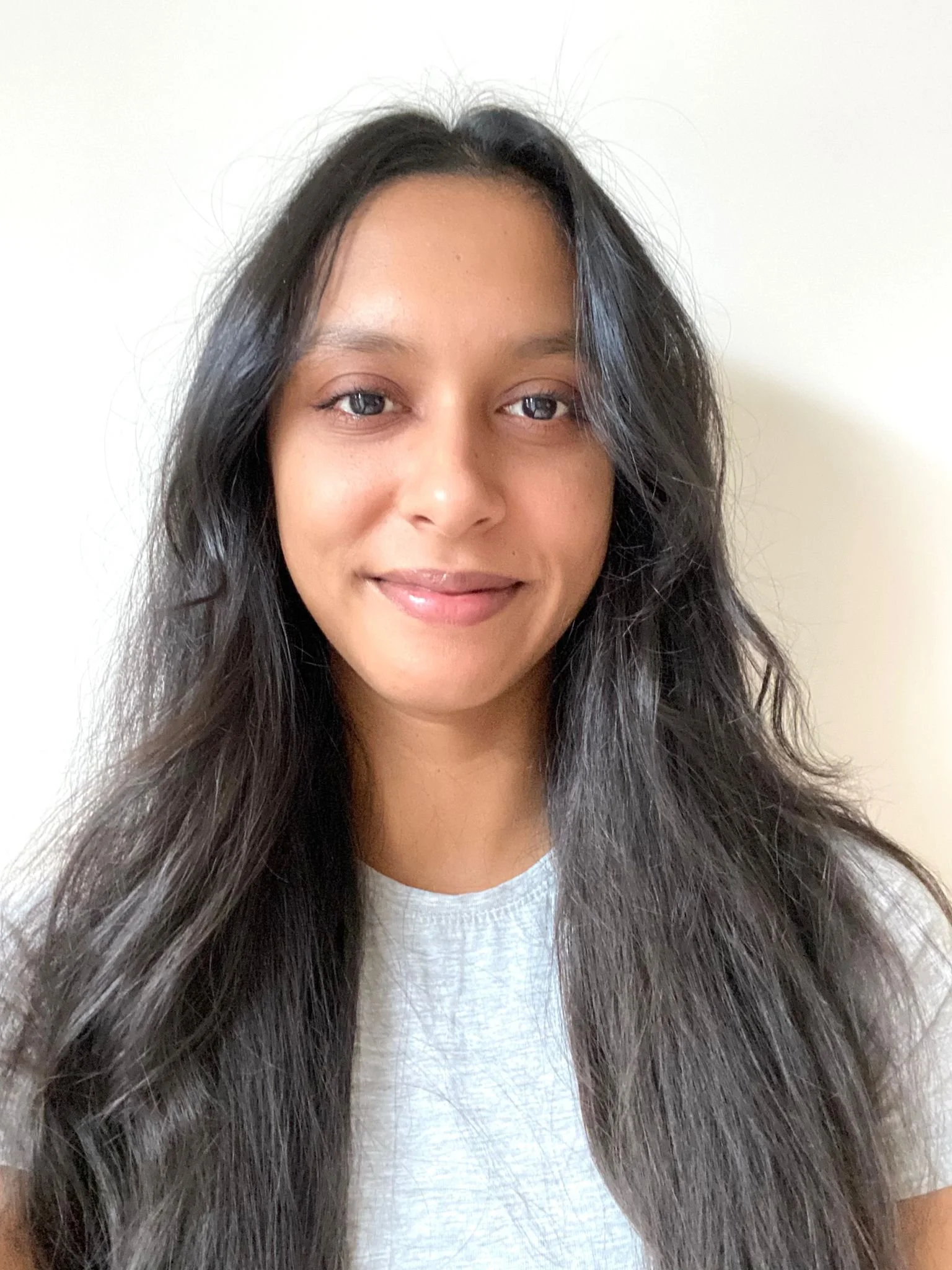 Close-up of a young woman with long wavy dark hair, dark skin, and brown eyes smiling slightly against a plain white wall background.