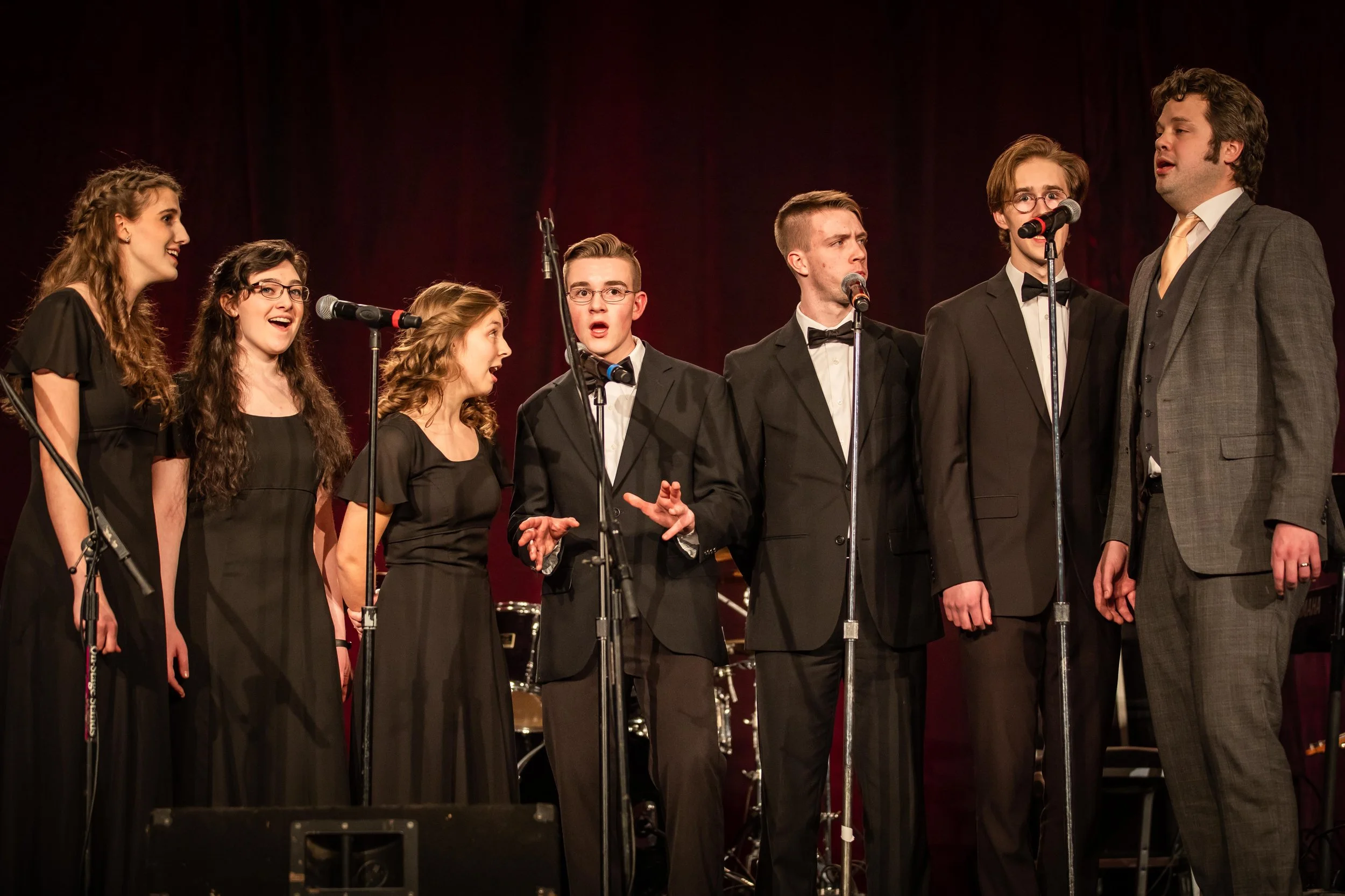 A diverse group of seven young singers on stage performing together. They are dressed in formal attire, with the women in black dresses and the men in black tuxedos with bow ties. Two microphones are in front of them, and they are singing passionately. The background is a dark red curtain, indicating a concert or recital setting.