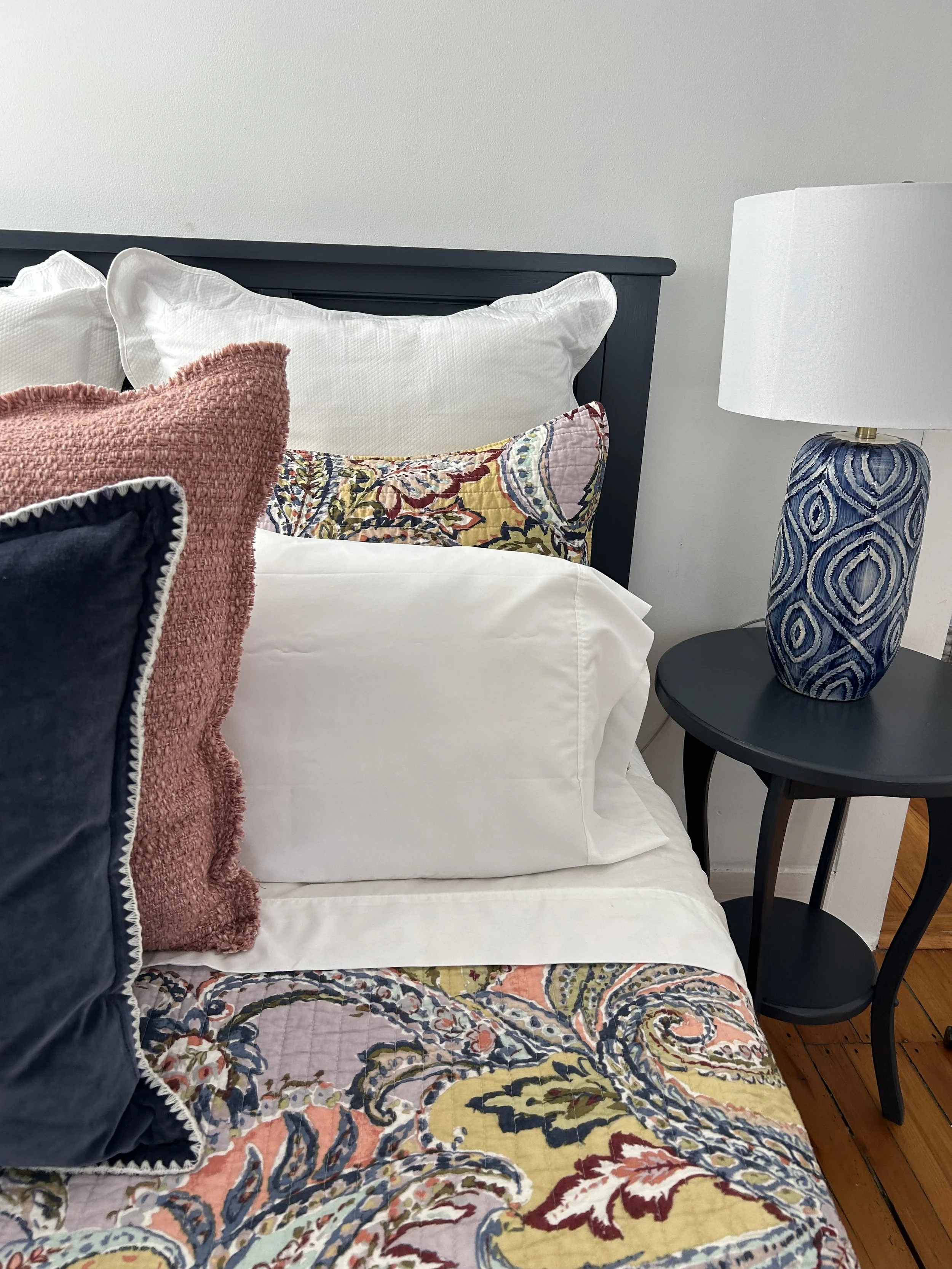 A bedroom with a blue and white patterned quilt, white pillows, a pink textured throw, and a black side table with a blue textured lamp. The bed has a black headboard and a plain white wall in the background.