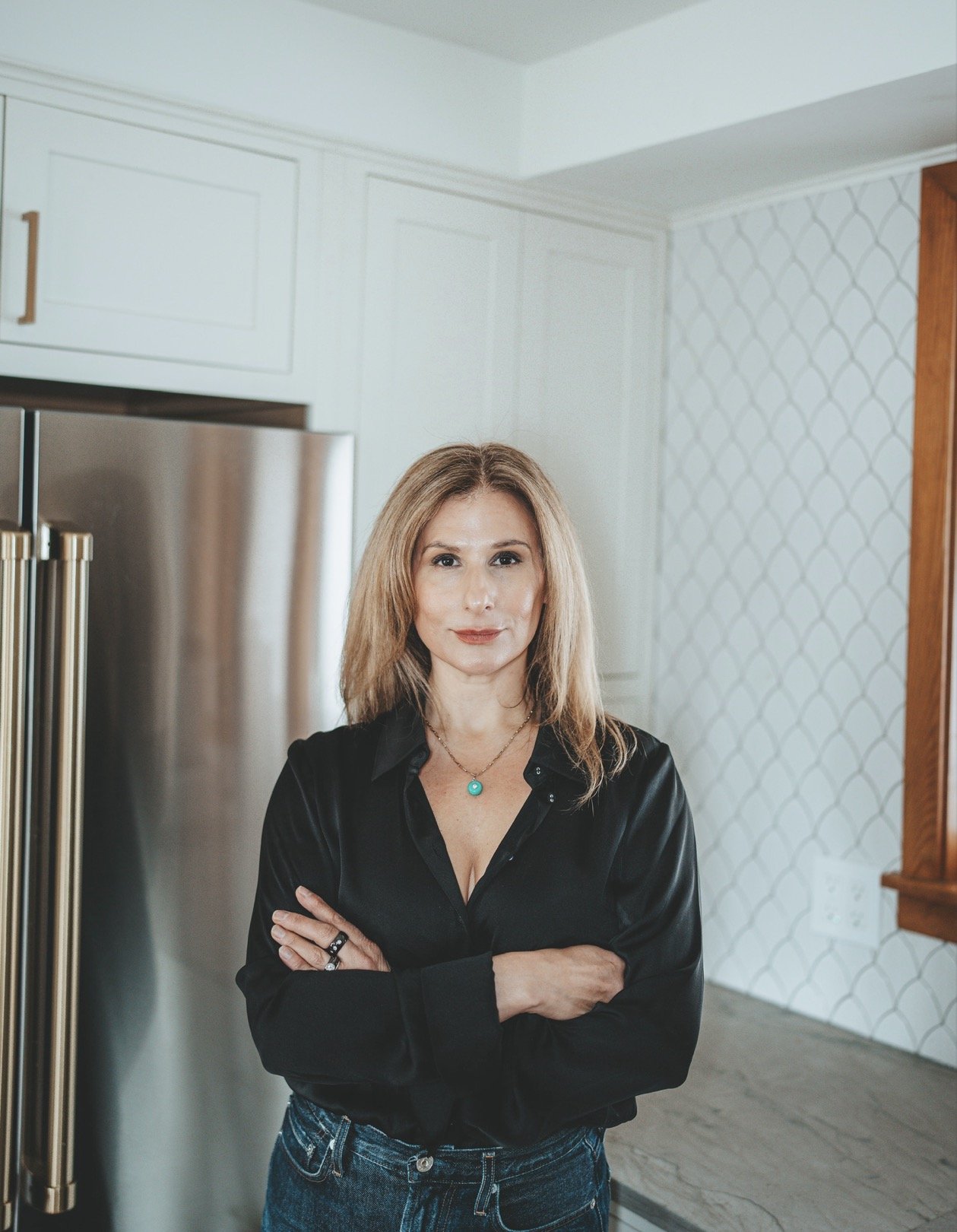 A woman with blonde hair and light skin standing in a kitchen, crossing her arms, wearing a black shirt and blue jeans, with a turquoise necklace.