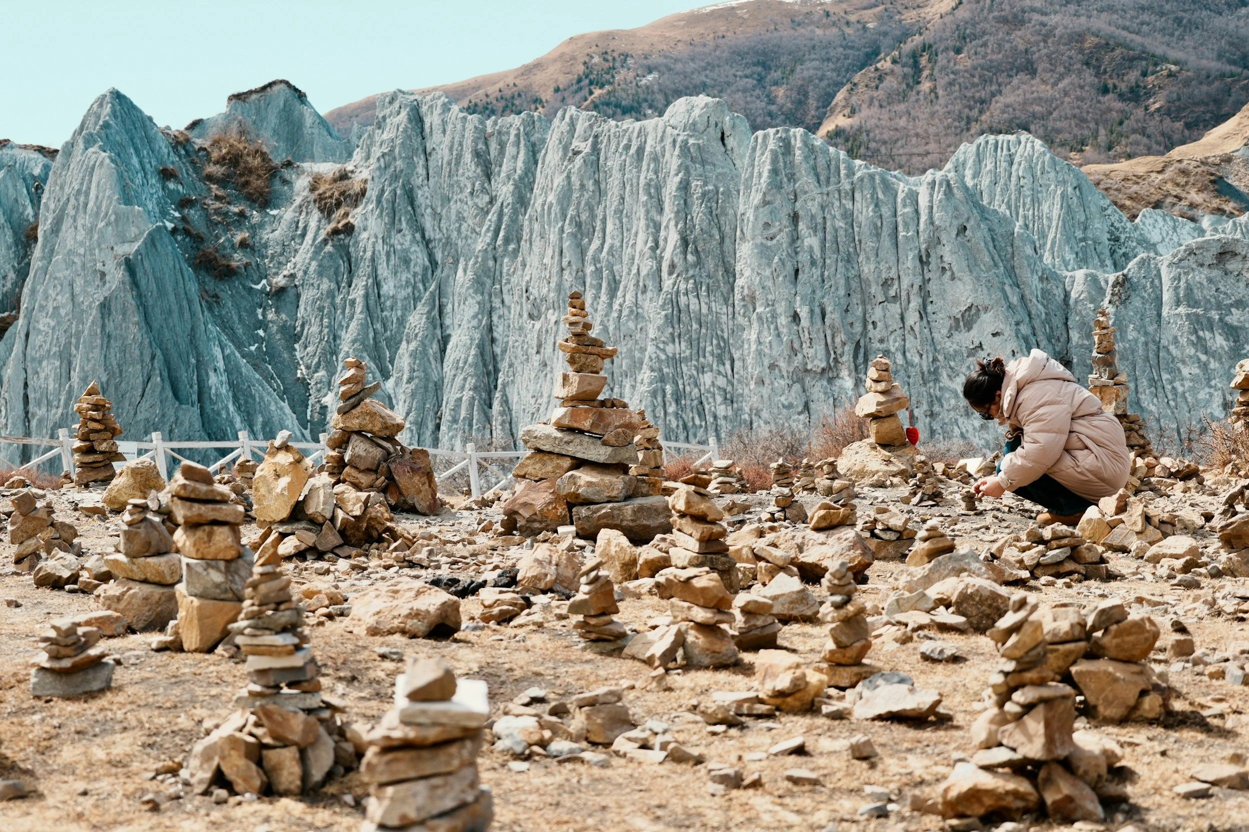 Person in a beige jacket arranging small rocks into stacked cairns in front of a glacier and mountain landscape.