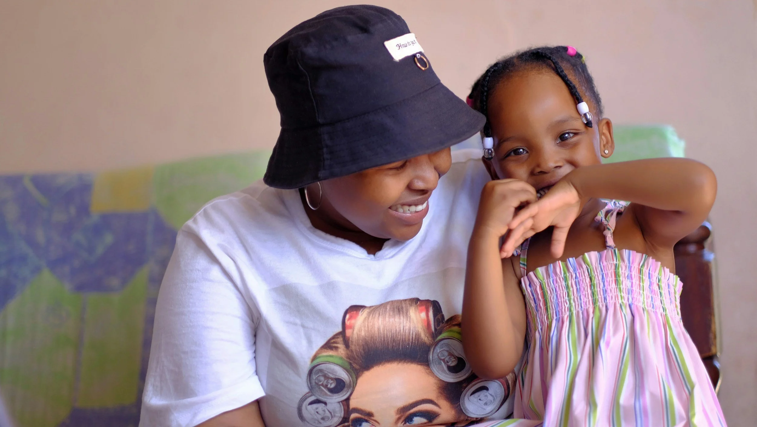 A woman wearing a black hat and white t-shirt is smiling and holding a young girl with black braided hair and colorful beads. The girl is wearing a striped dress with pink, green, and yellow colors, and is smiling with her arm raised near her face. They are sitting against a background of a painted wall with an abstract pattern.