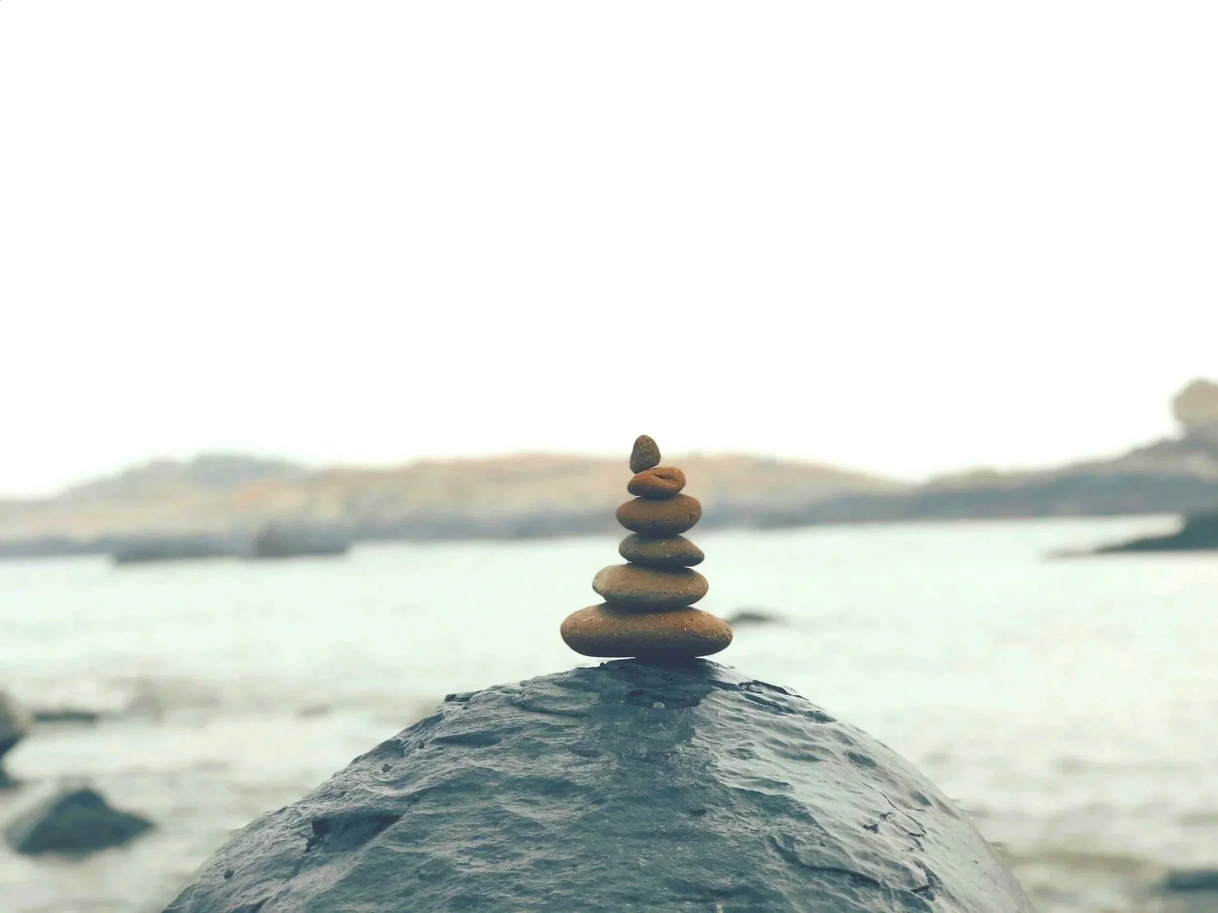 Stacked stones balanced on a rocky surface near a body of water against a cloudy sky background.