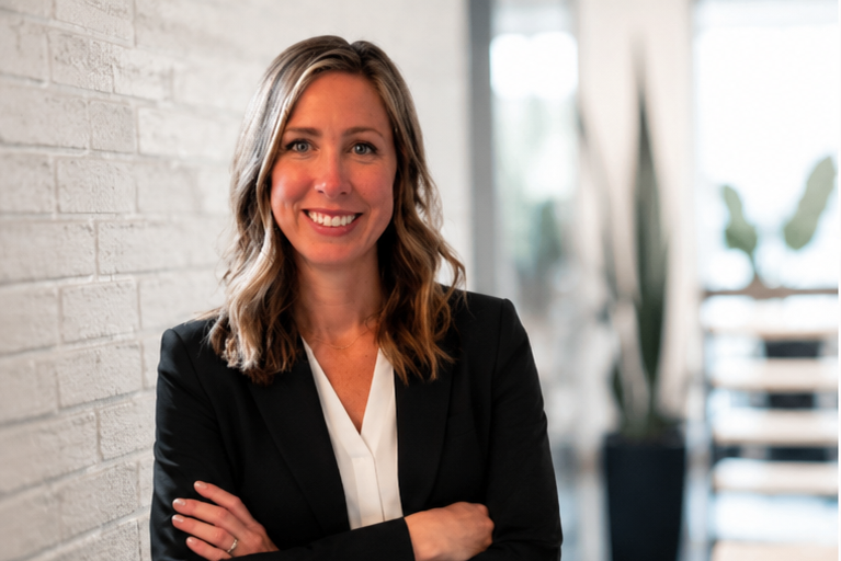 A woman with shoulder-length wavy hair is smiling and standing with arms crossed inside an office. She is wearing a black blazer over a white blouse, and the background features a white brick wall and large windows with plants outside.