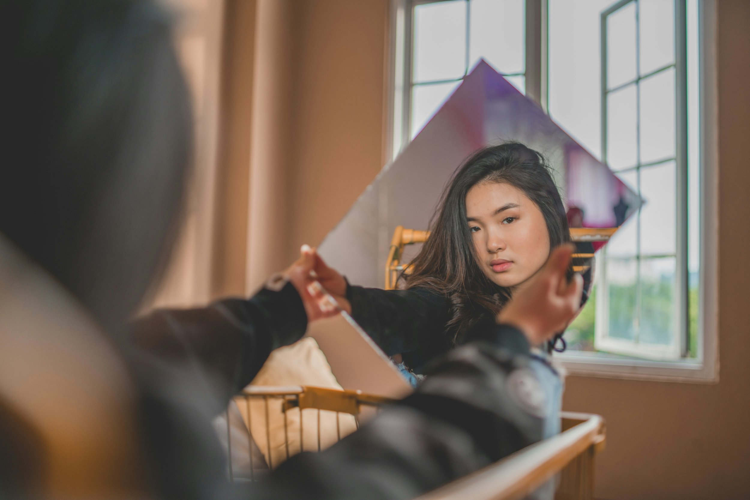 A woman with long dark hair is looking into a mirror, adjusting or applying makeup, with a window and natural light behind her.