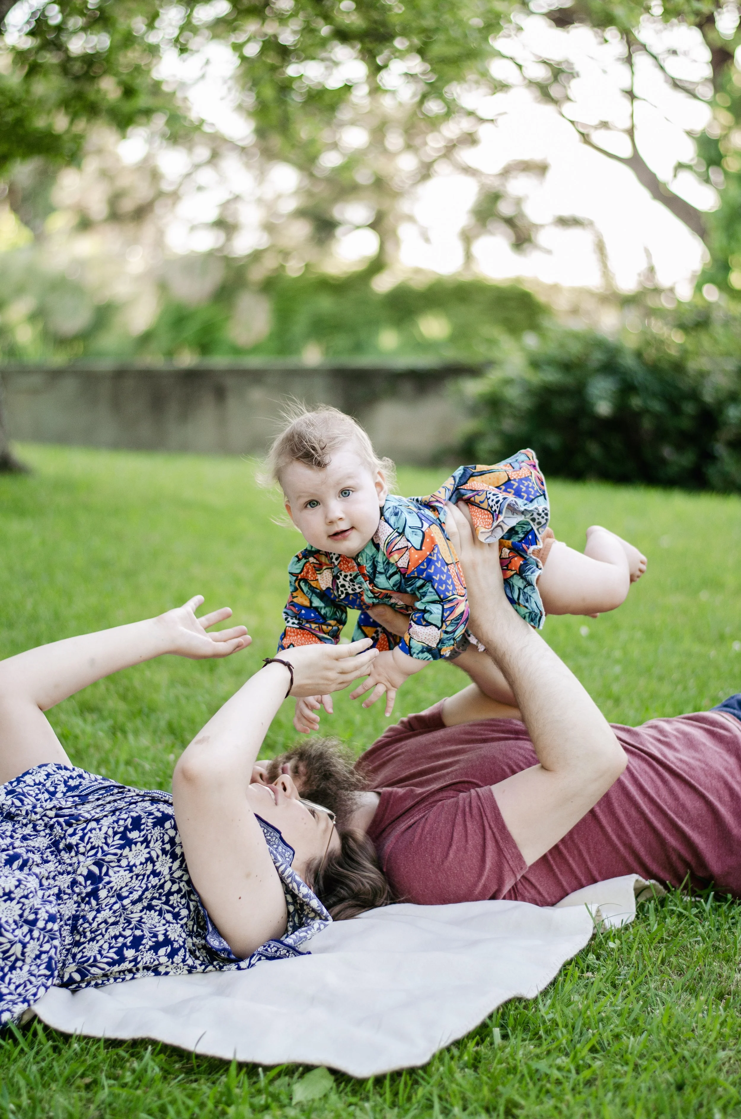 A woman lying on a blanket on the grass outdoors, holding a young child above her. The child is reaching out with a hand and looking toward the camera. The background shows greenery and trees with sunlight filtering through.