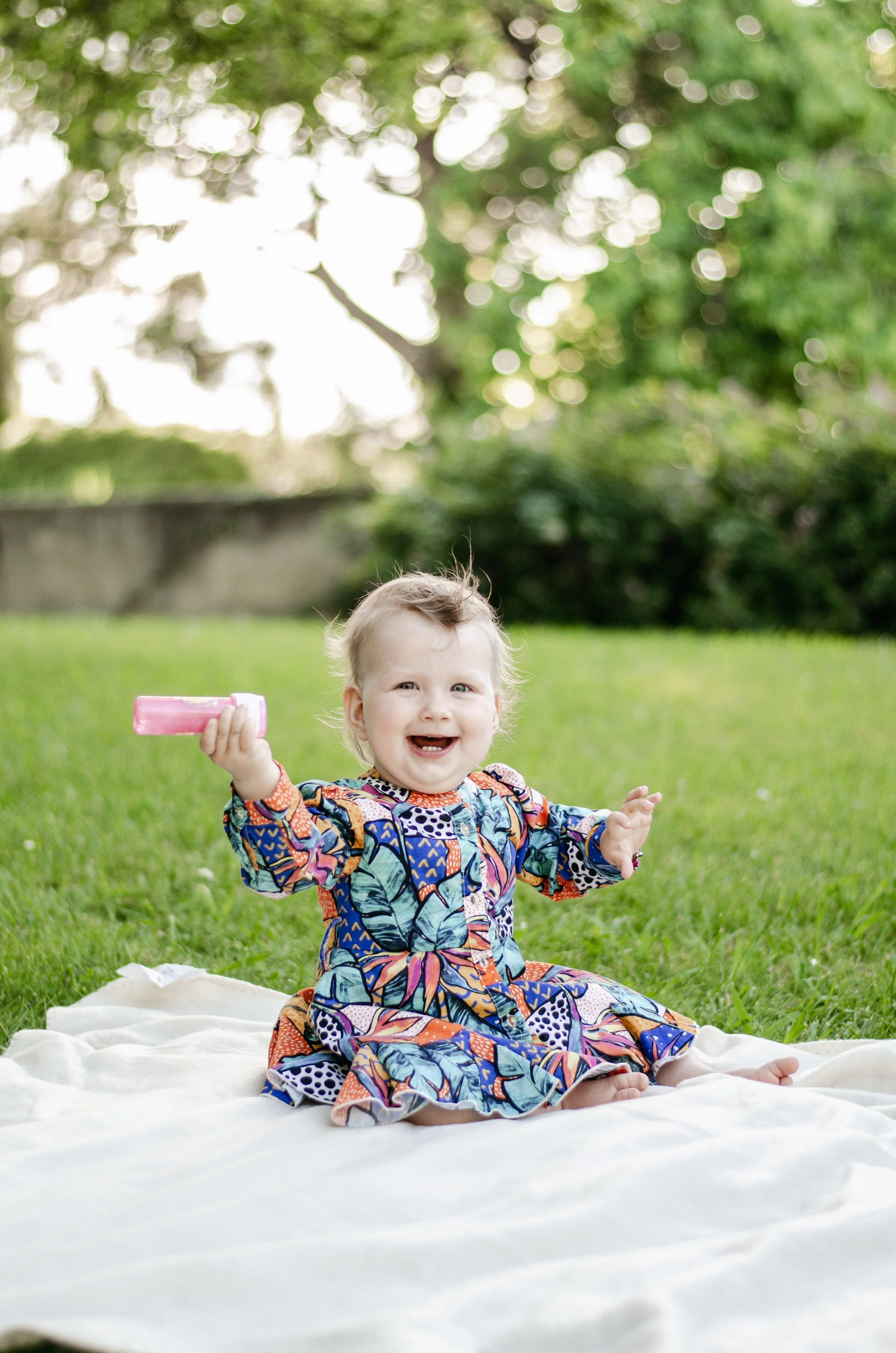A smiling toddler with curly blonde hair, wearing a colorful patterned dress, sitting on a white blanket on the grass outdoors. The child is holding a pink toy or object in one hand and has arms outstretched. The background features green trees and s