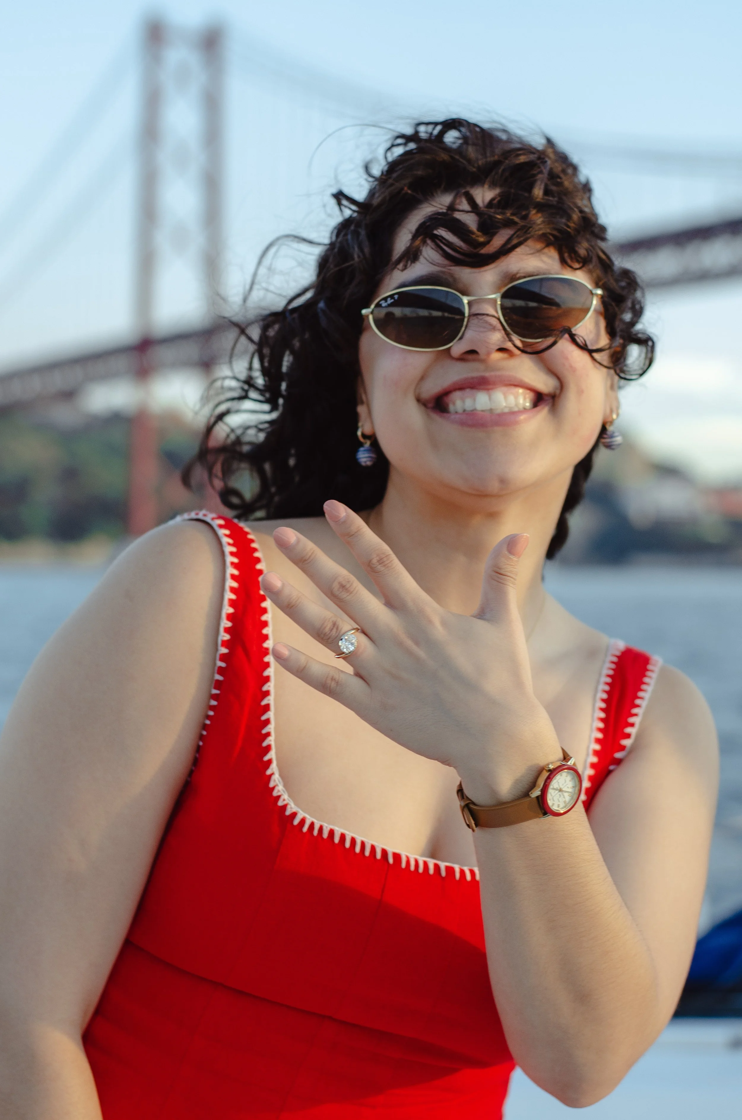 A woman smiling outdoors with sunglasses and a red dress, with a bridge in the background.