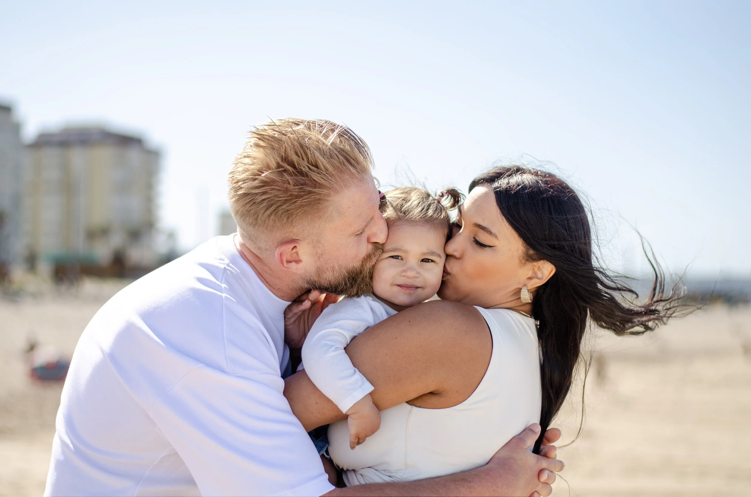 A happy family of three, a man, woman, and a young girl, sharing a loving moment outdoors on a sunny day, with the man and woman giving kisses to the girl.