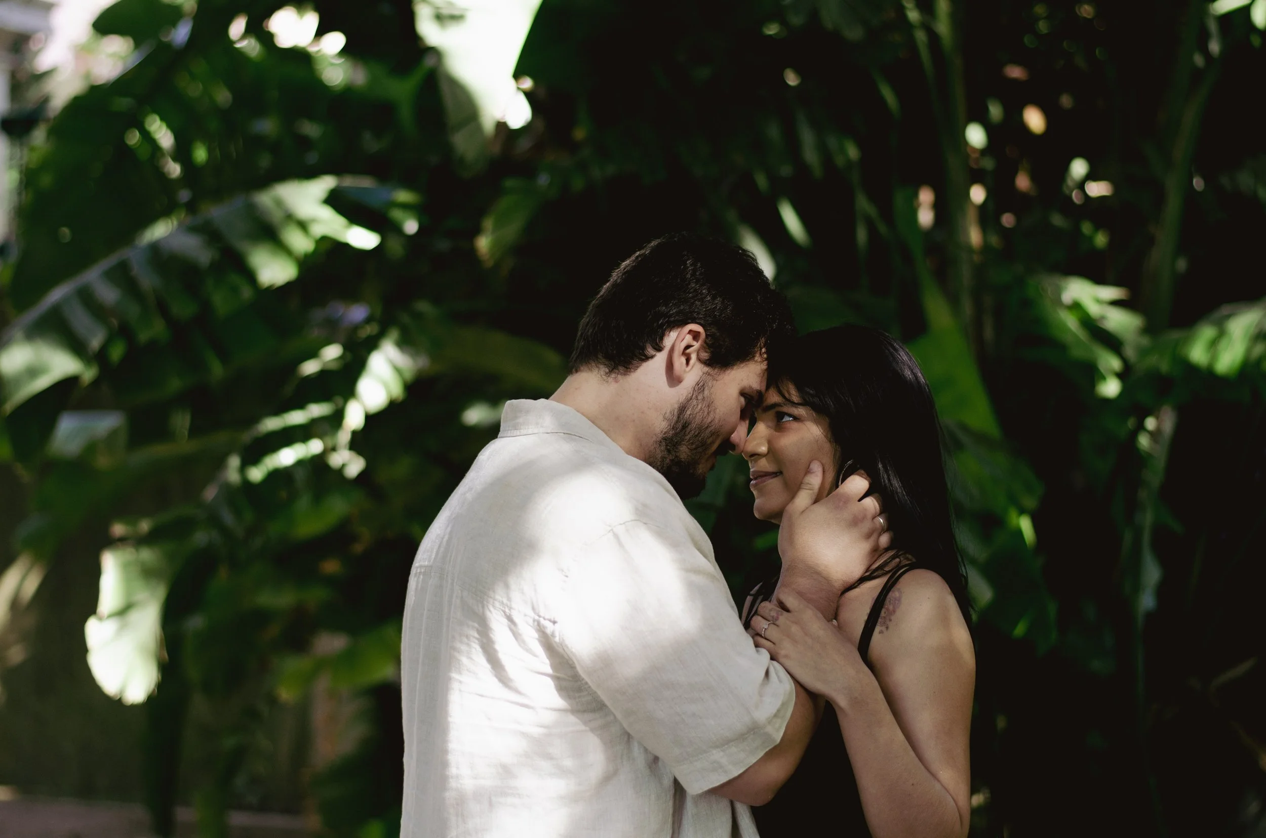 A couple is embracing affectionately in a lush green outdoor setting, with both their eyes closed and foreheads touching.