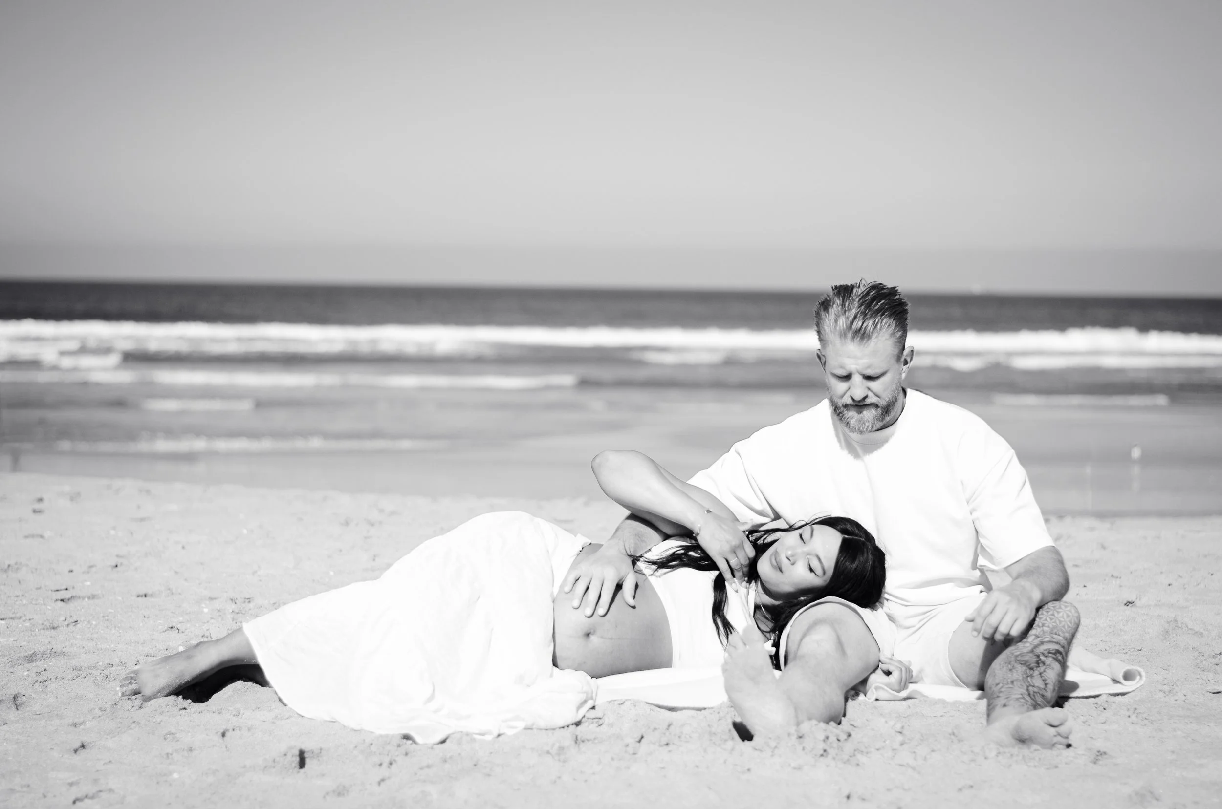 A man with a beard and a woman with long dark hair lying on the sand at the beach, with the man giving the woman a massage or physiotherapy treatment. The ocean and sky are visible in the background.