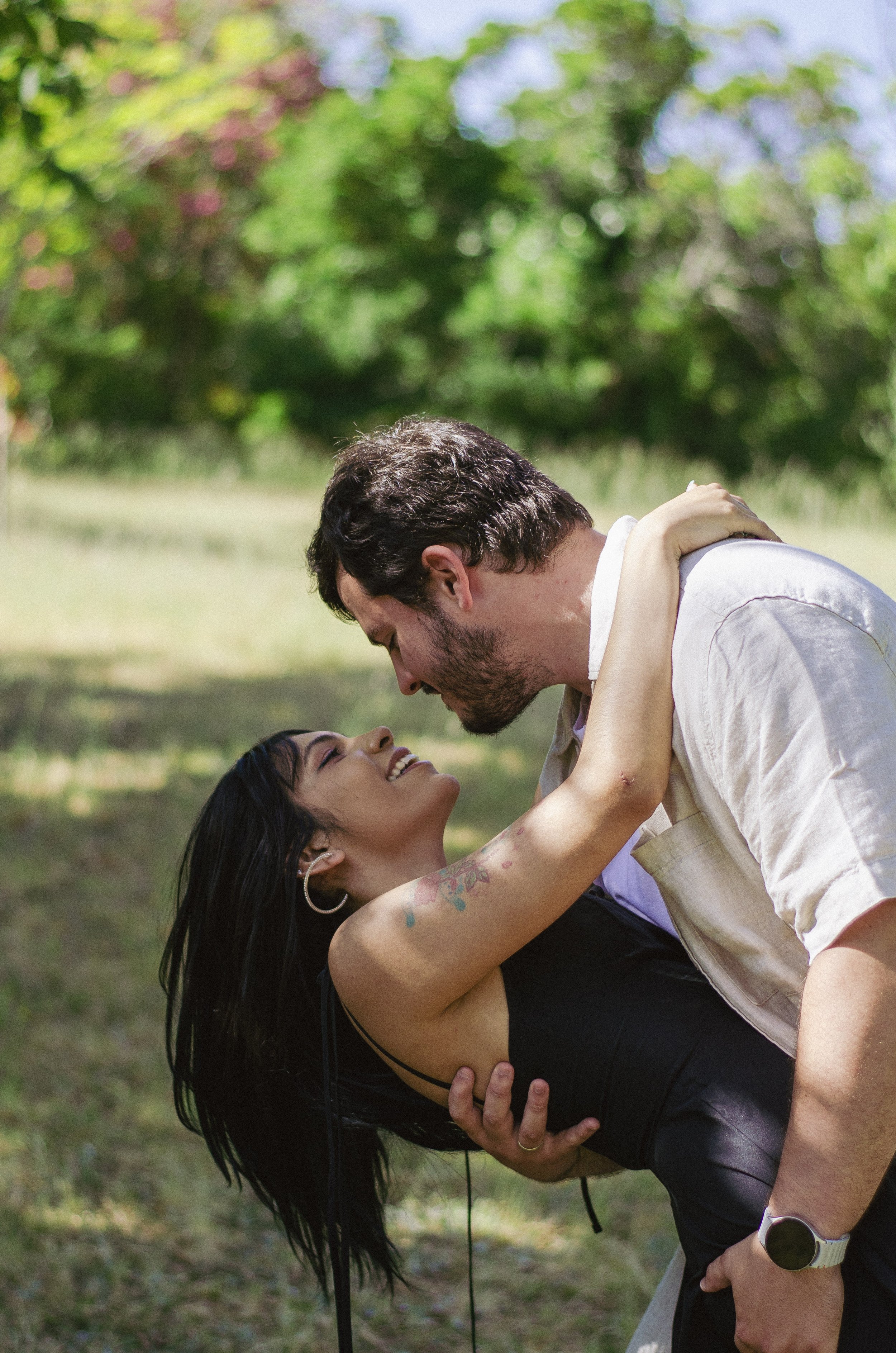 A man and woman are outdoors in a green, leafy area. The woman has long dark hair and tattoos on her arm. They are smiling and leaning close, with the man holding the woman, and they appear to be happy and affectionate.