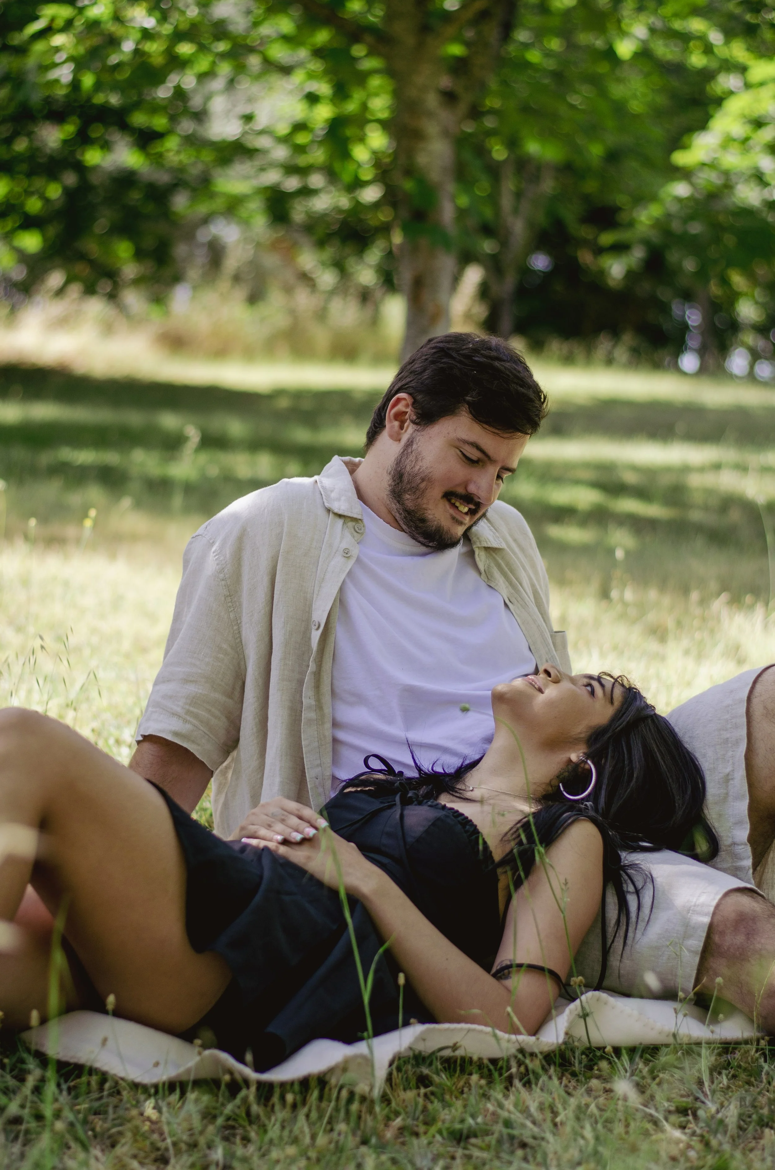 A man and woman lie on the grass outdoors, with the woman lying down and the man sitting beside her. The woman has dark hair and is wearing a black outfit. The man is wearing a light-colored shirt and has short dark hair and a beard. They are surroun