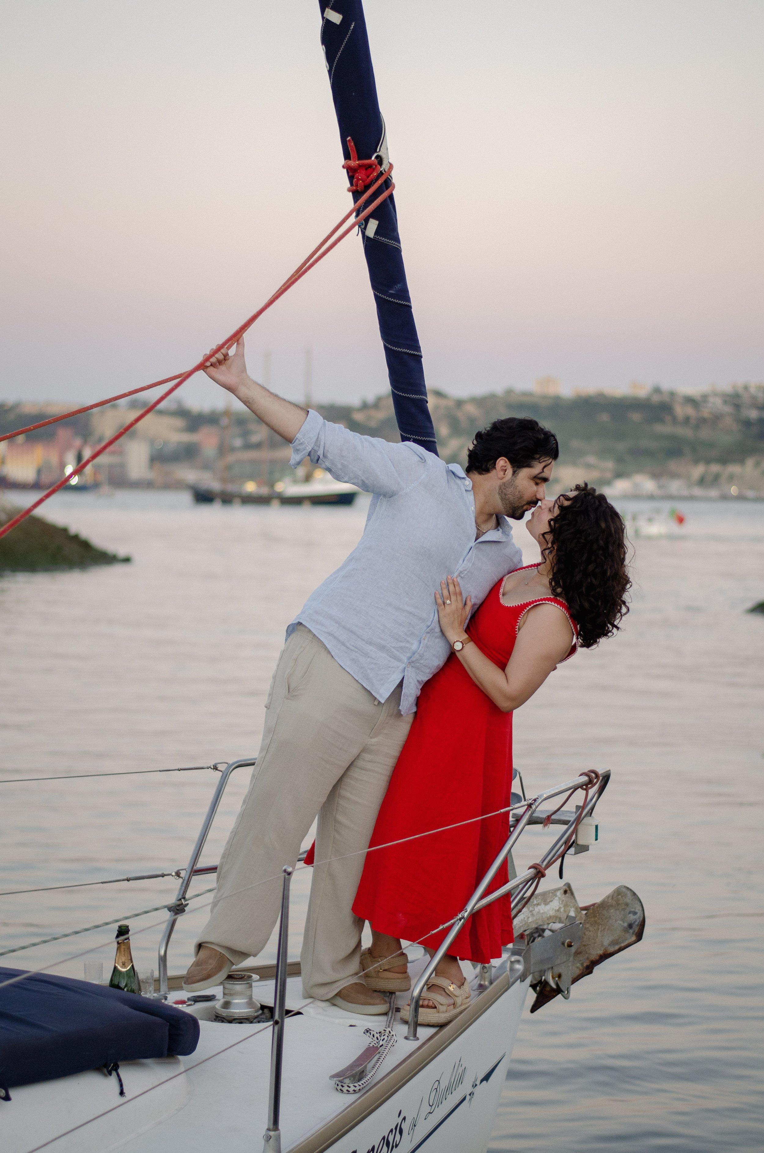 A man and woman on a sailboat, embracing and about to kiss, with a harbor and city buildings in the background during sunset.