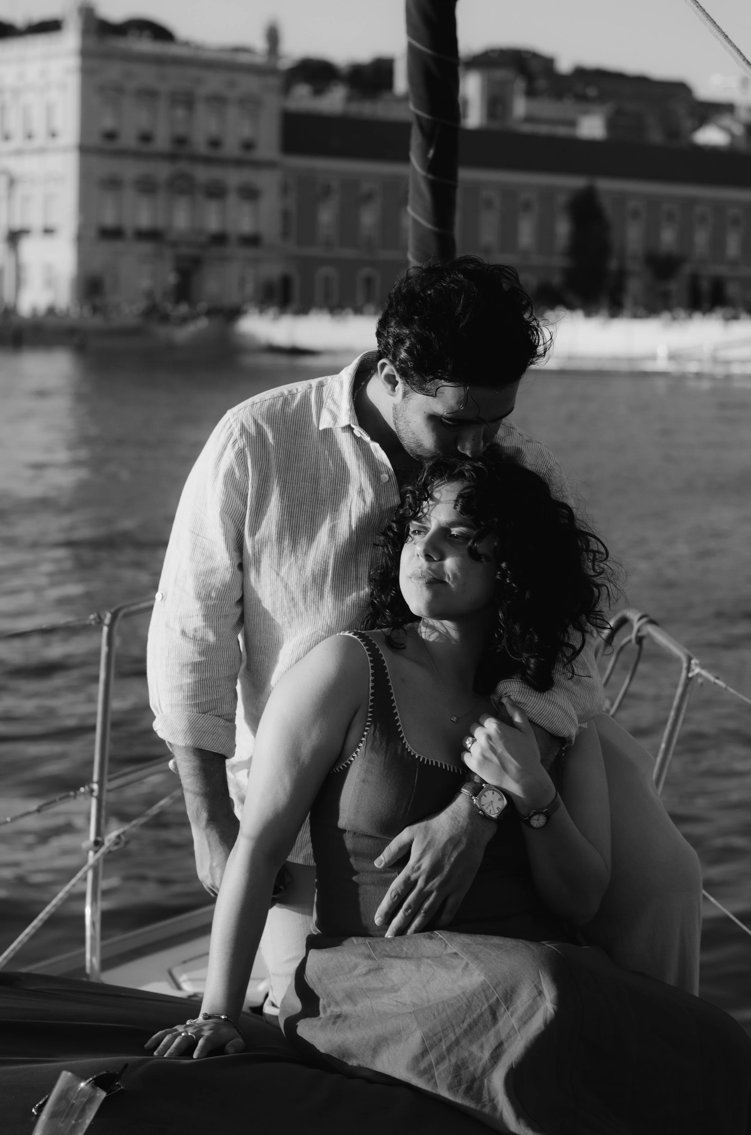 A black and white photo of a man and woman on a boat. The man with dark curly hair is leaning over the woman, who has curly hair and is wearing a sleeveless top. The woman is seated, holding onto her chest as the man leans in close. In the background