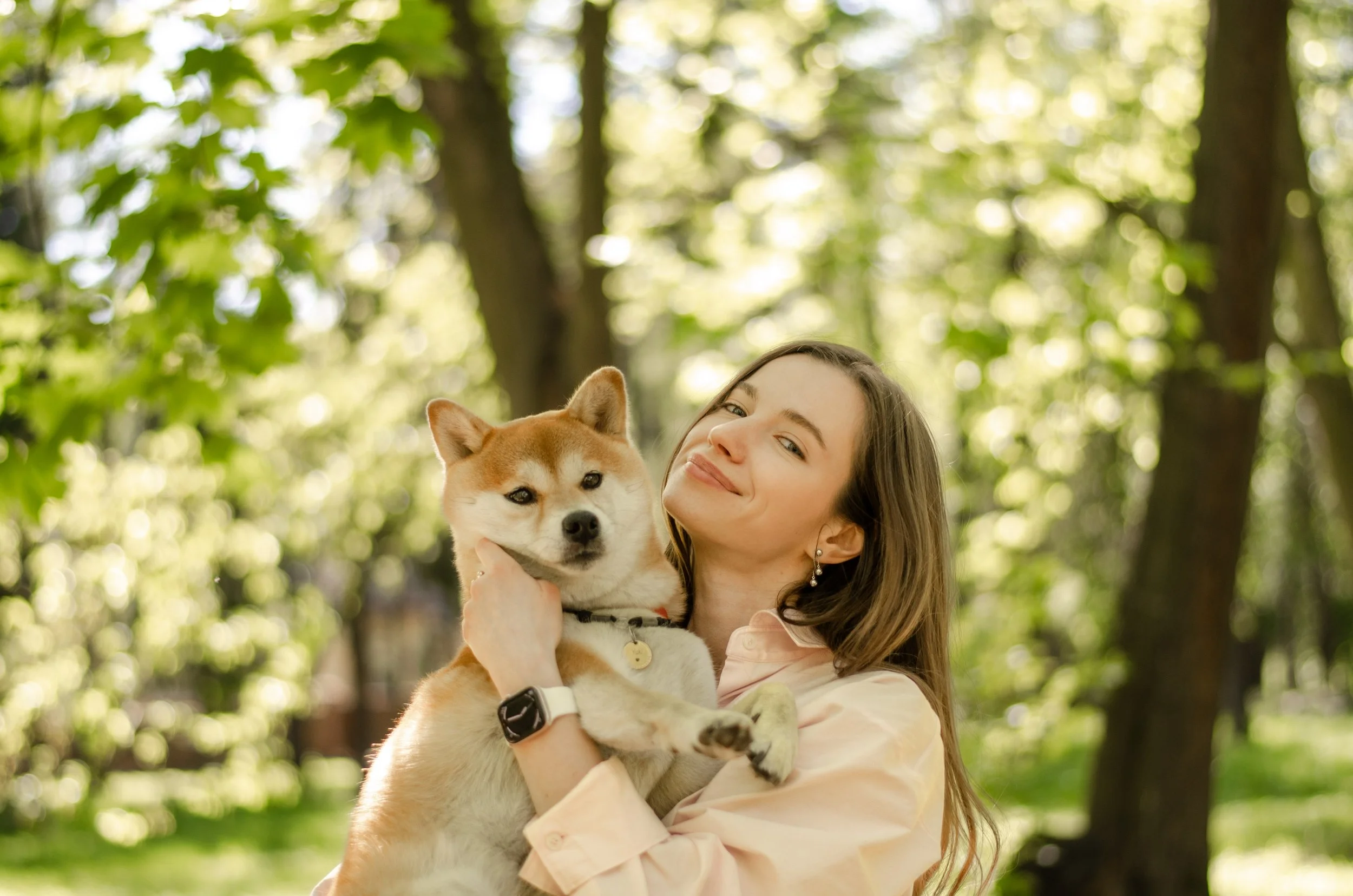 A young woman smiling while holding a Shiba Inu dog outdoors in a wooded area with sunlight filtering through green leaves.