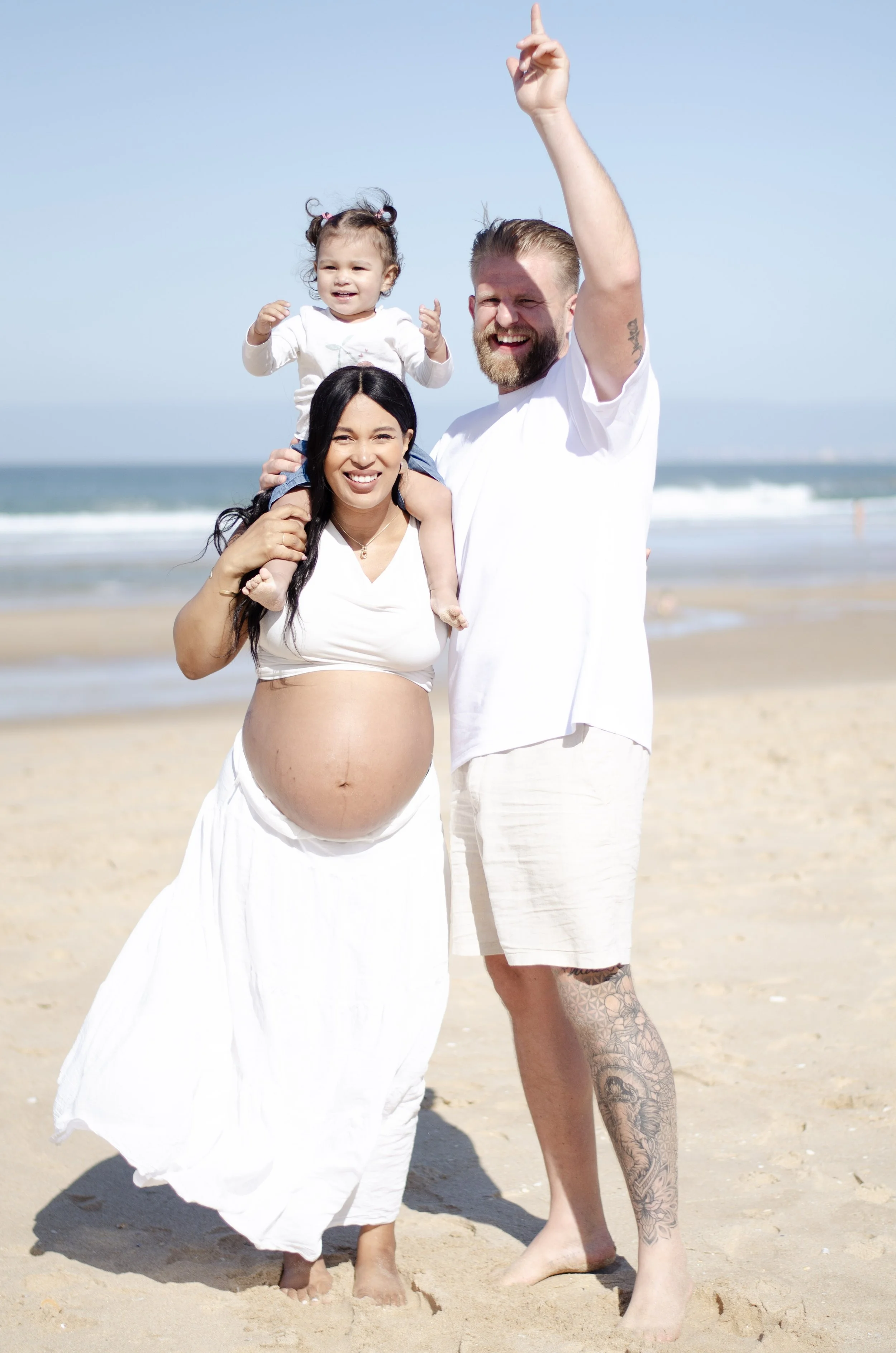A pregnant woman, a man, and a young girl are on the beach. The woman is holding the girl on her shoulders, all smiling and wearing white clothing. The man is standing next to them, raising his hand with one finger pointing up, with a beard and tatto