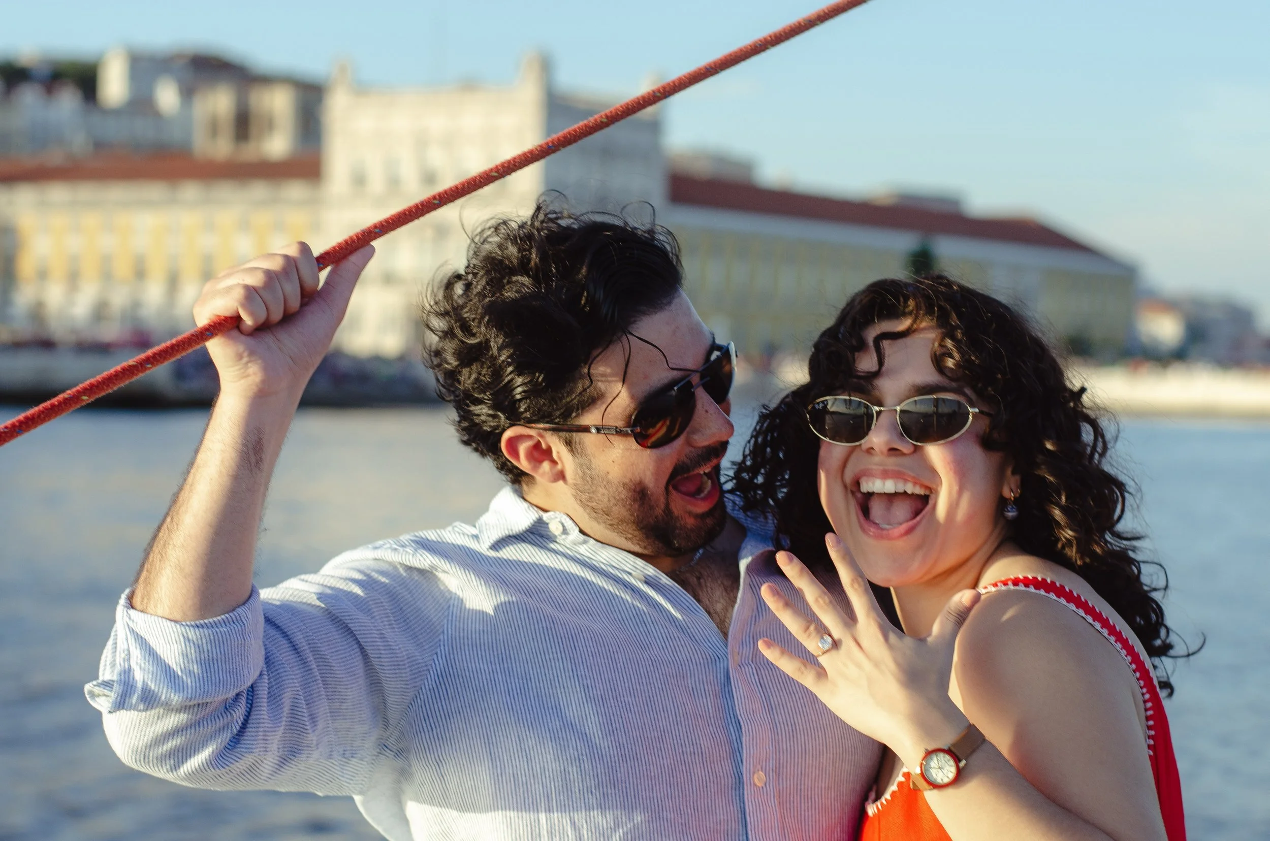 A happy couple taking a selfie outdoors near water, with buildings in the background, both wearing sunglasses and smiling.