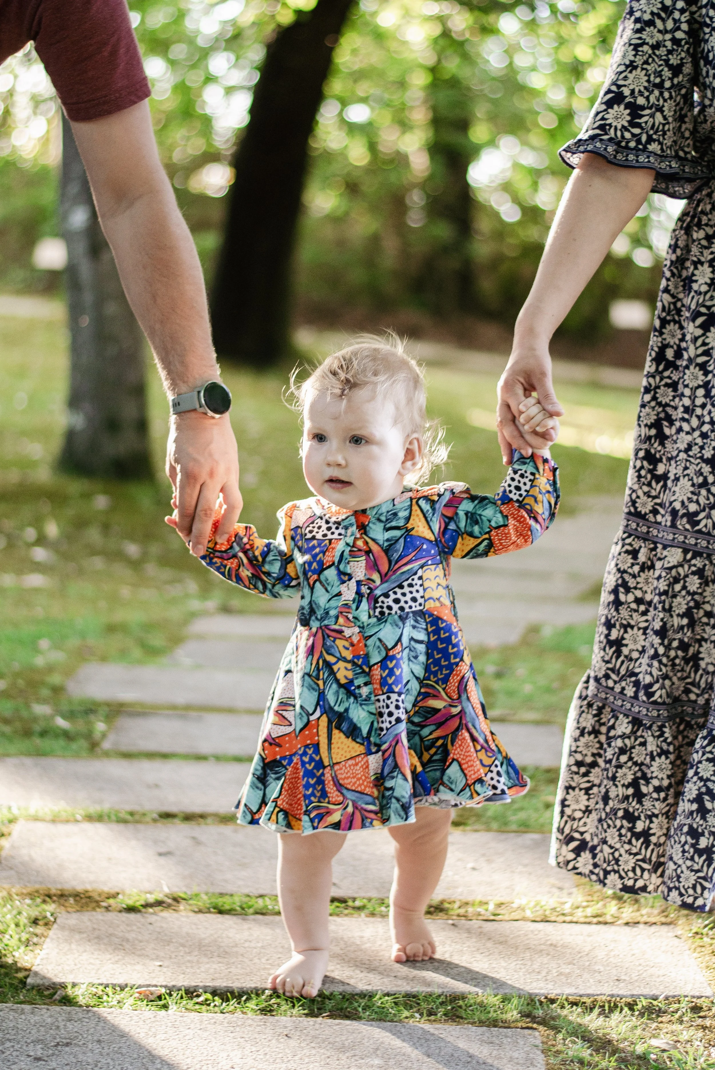 A young girl with curly blonde hair wearing a colorful patterned dress walking outside on a sidewalk, holding hands with an adult woman dressed in a floral dress. The scene is set in a green park with sunlight filtering through trees.