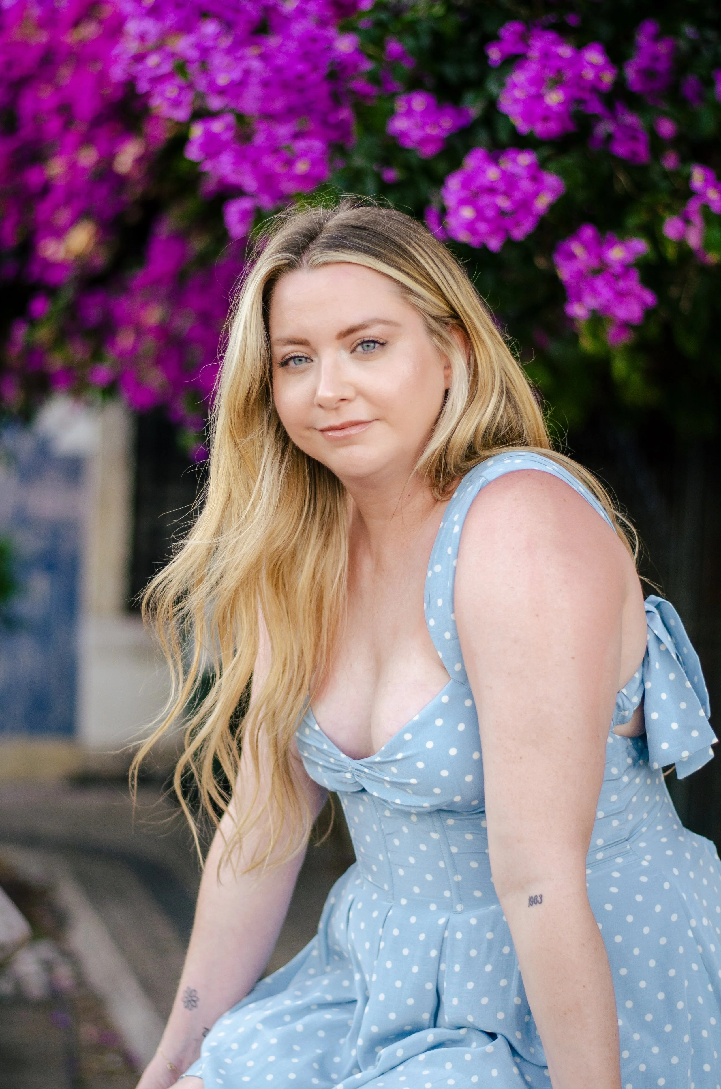 A young woman with long blonde hair and blue eyes wearing a blue polka dot sundress with ruffled sleeves, sitting outdoors in front of purple flowers.