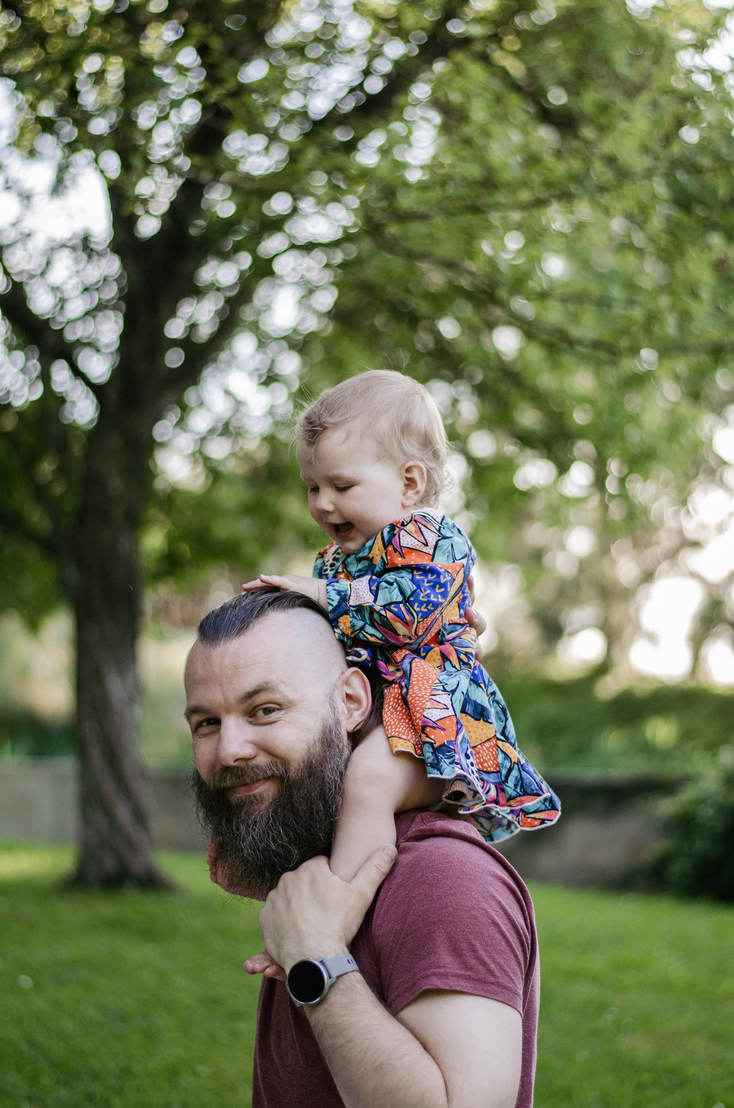 A man with a beard is giving a piggyback ride to a young boy with curly hair and colorful clothes, outdoors in a park with green grass and trees in the background.