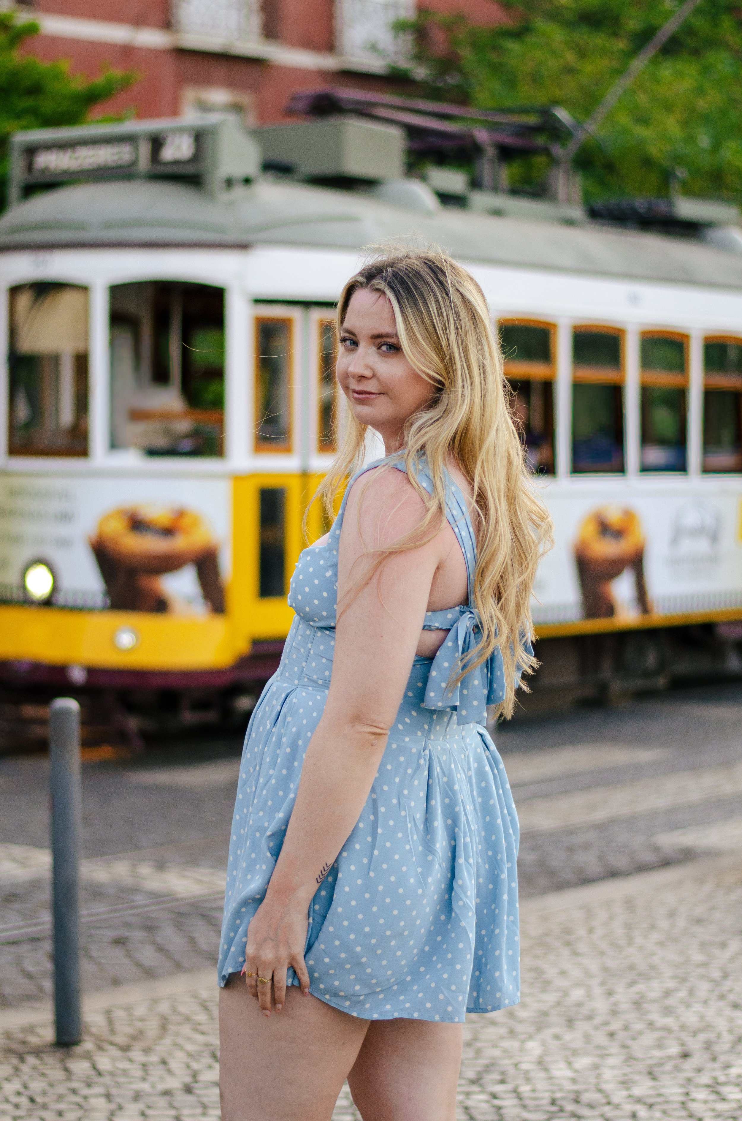 A blonde woman in a blue polka dot dress standing outside in front of a white and yellow trolley bus with a red brick building and green trees in the background.