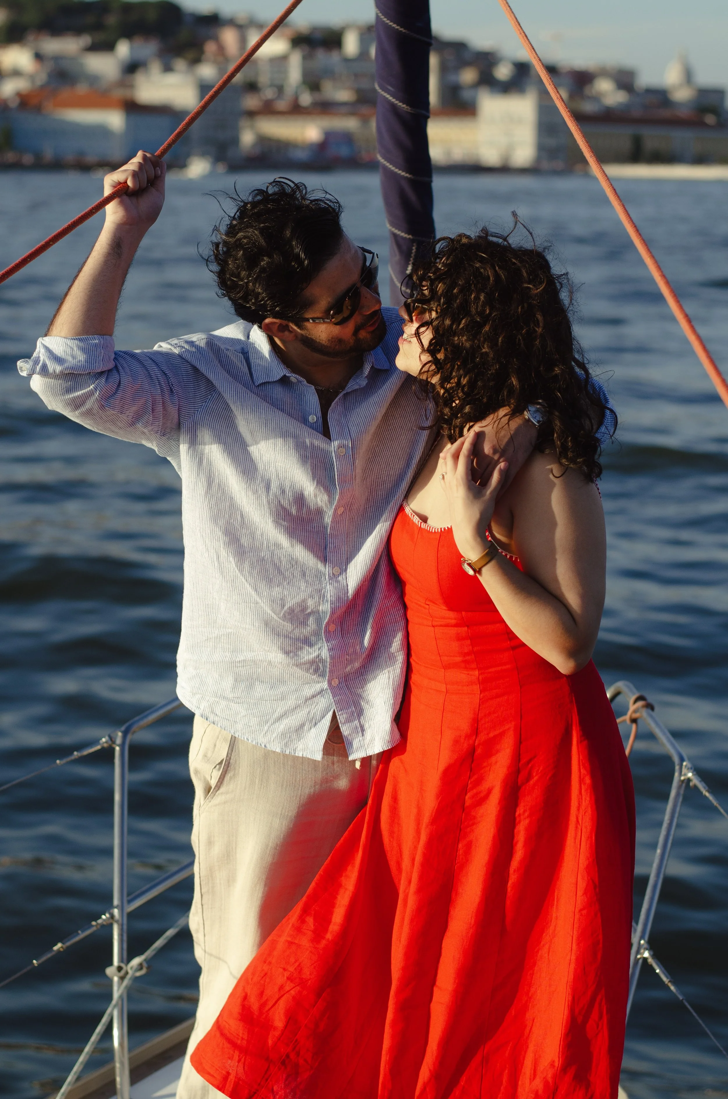 A man and woman on a sailboat during sunset, embracing and gazing into each other's eyes with a city skyline in the background.