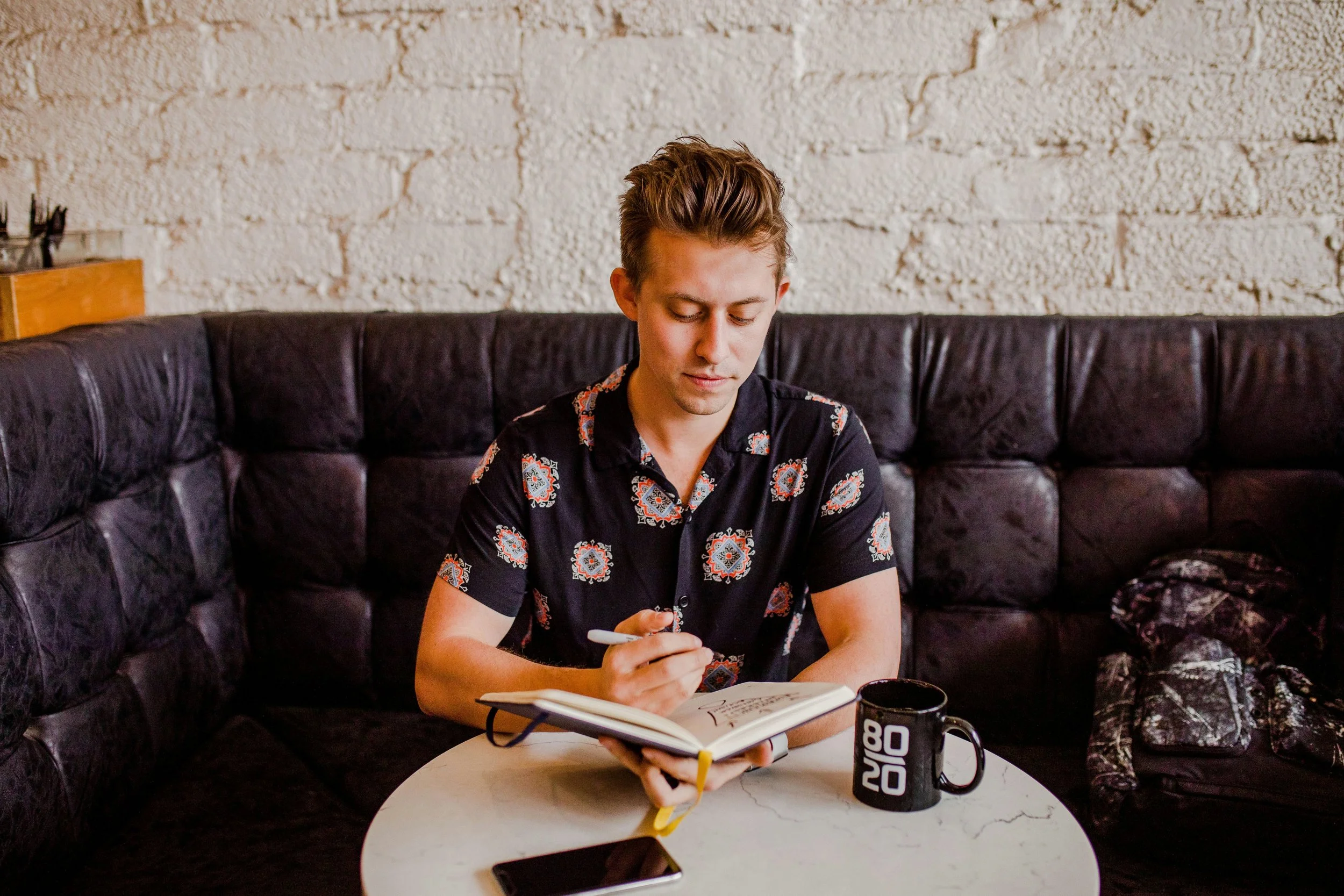 Young man with styled hair wearing a patterned black shirt, sitting on a black leather couch in a cafe, writing in a notebook on a white marble table with a black coffee mug and smartphone.