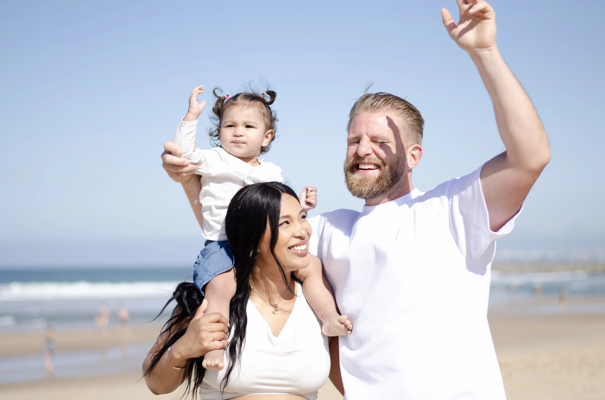A happy family enjoying a day at the beach, with a man holding a young girl on his shoulders and a woman beside them smiling, all wearing casual white clothing under a clear blue sky.