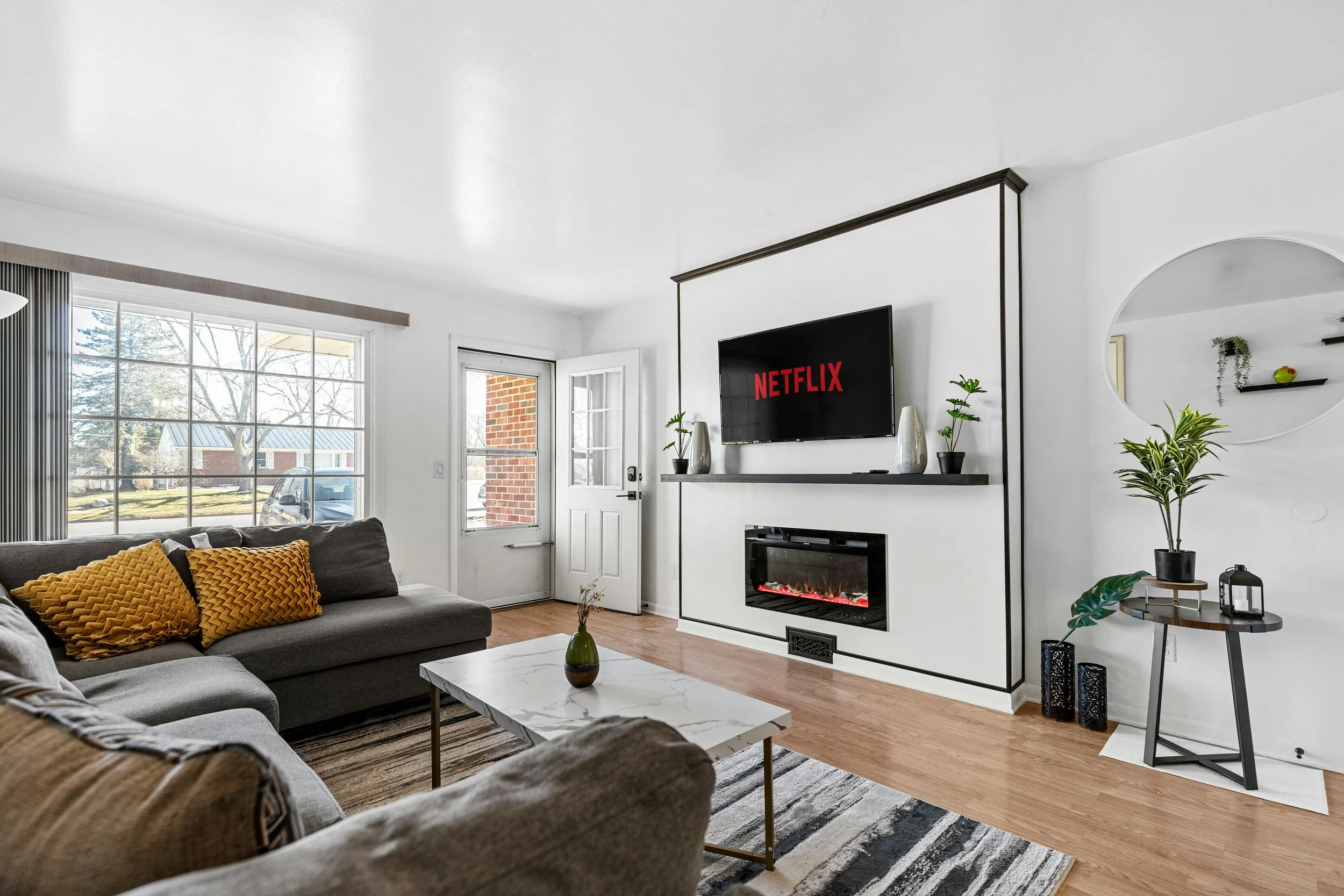 Living room with large window, sliding glass door, gray sofa with yellow pillows, white marble coffee table, wall-mounted TV displaying Netflix, electric fireplace, and indoor plants.