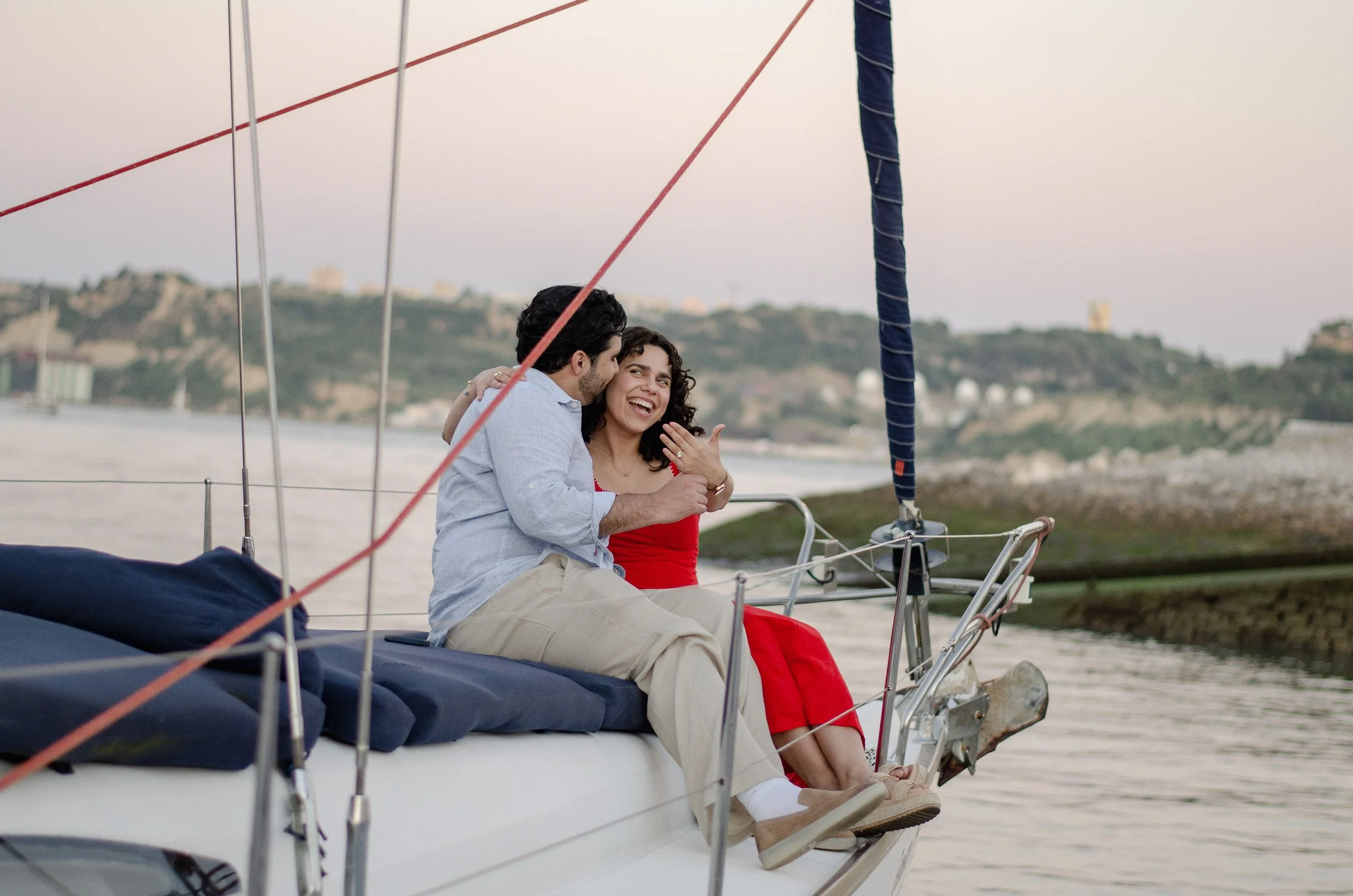 A couple sitting on a sailboat, smiling and enjoying each other's company near the water with a distant shoreline visible in the background.