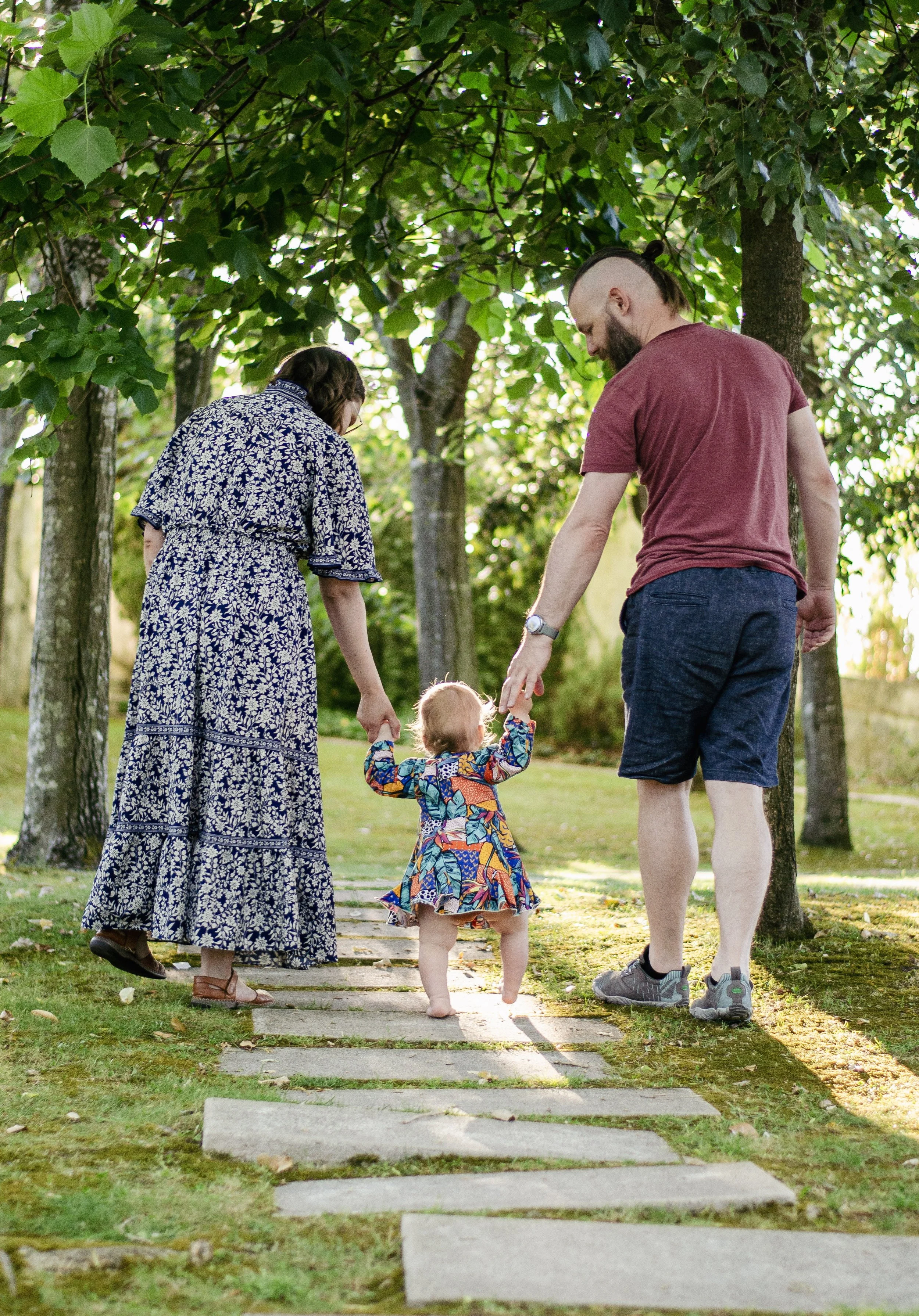 A family of three walking hand in hand on a sidewalk through a park, surrounded by green trees. The woman wears a long dress with a floral pattern, the man wears a red t-shirt and shorts, and the child wears a colorful dress.