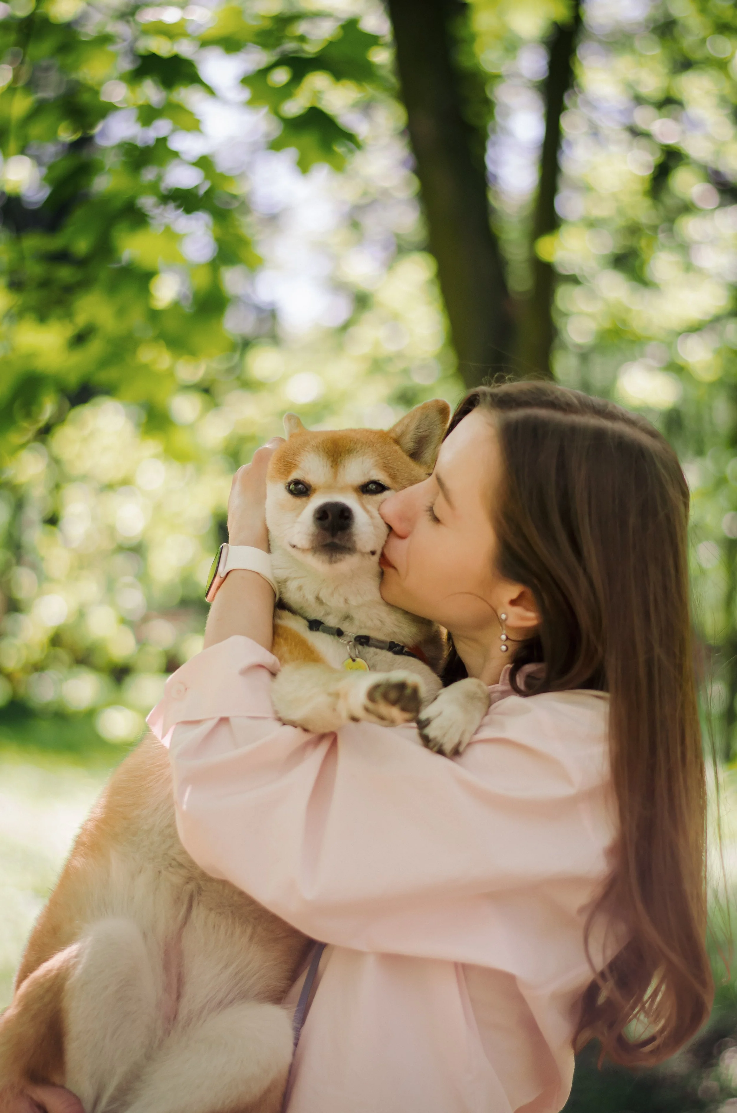 A woman with long brown hair, wearing a pink blouse, kisses a large, tan and white Shiba Inu dog on the face while holding it close outdoors in a wooded area with green leaves and sunlight filtering through the trees.