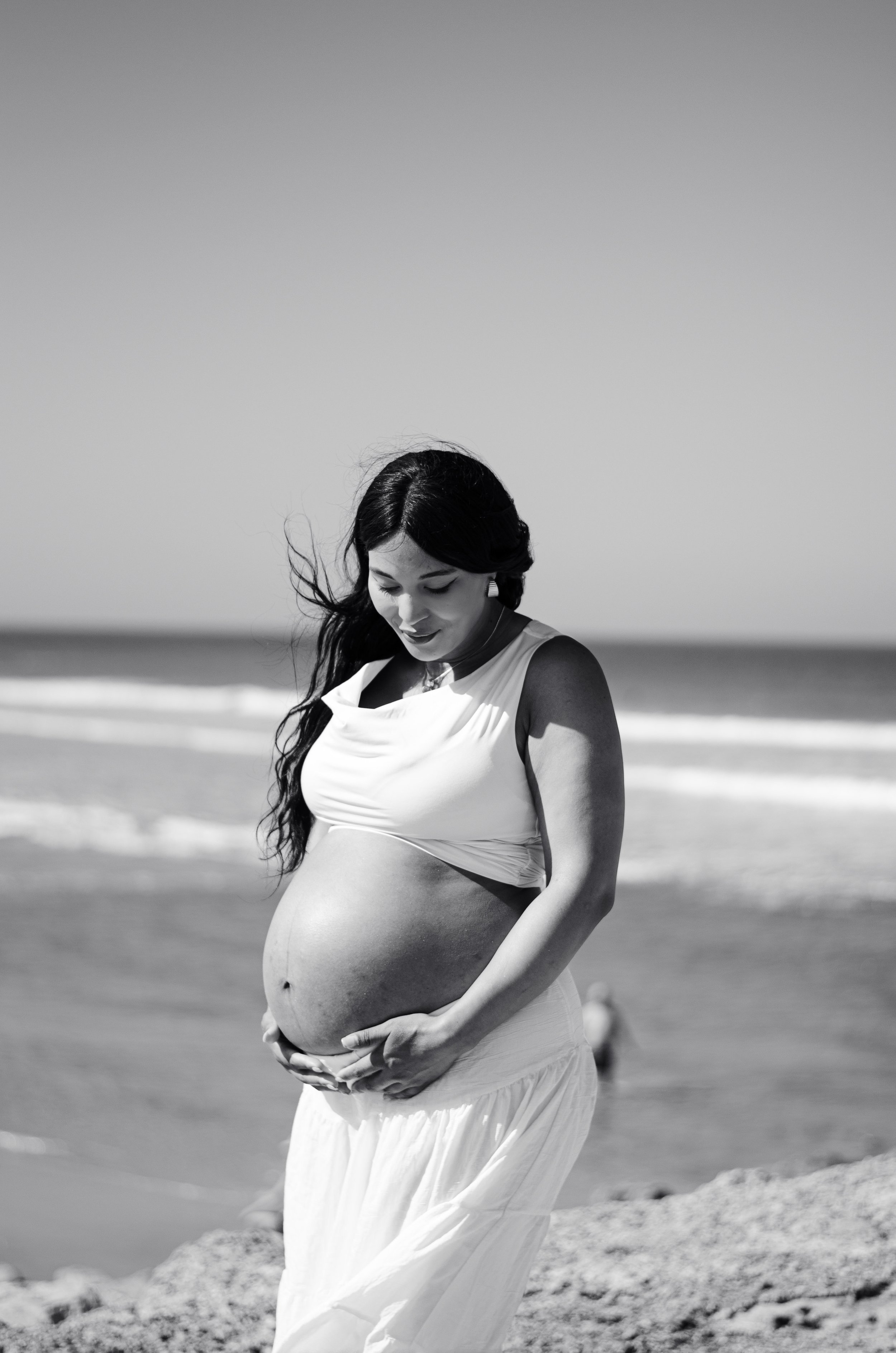 A pregnant woman on the beach, wearing a white dress, looking down at her belly with her hands gently cradling it, with wind blowing through her hair.