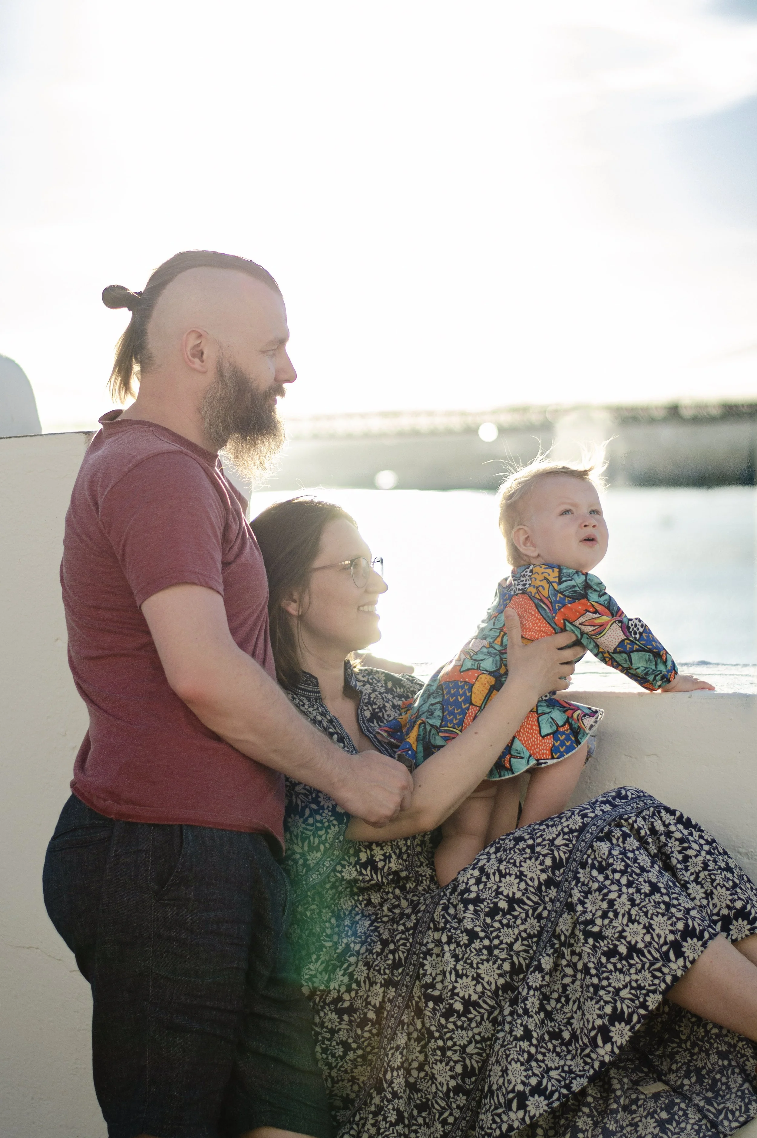 A group of four people, including a man with a beard and man bun, a woman with glasses, a child with spiky hair, and a woman with long hair, are enjoying a moment together outdoors by the water, with bright sunlight illuminating the scene.
