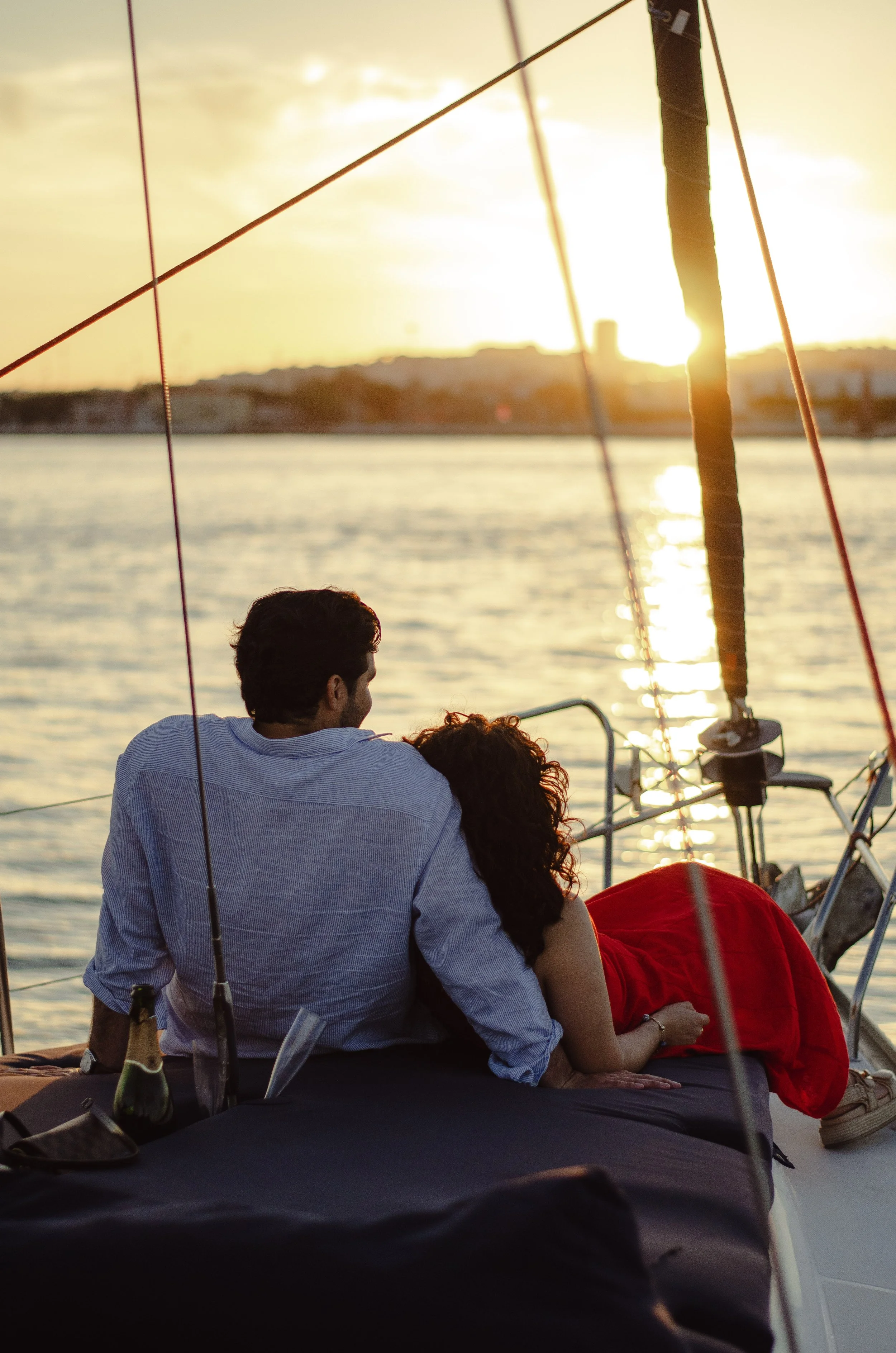Couple lying on a boat at sunset, facing the water, with the sun setting over the horizon, and sailing equipment nearby.