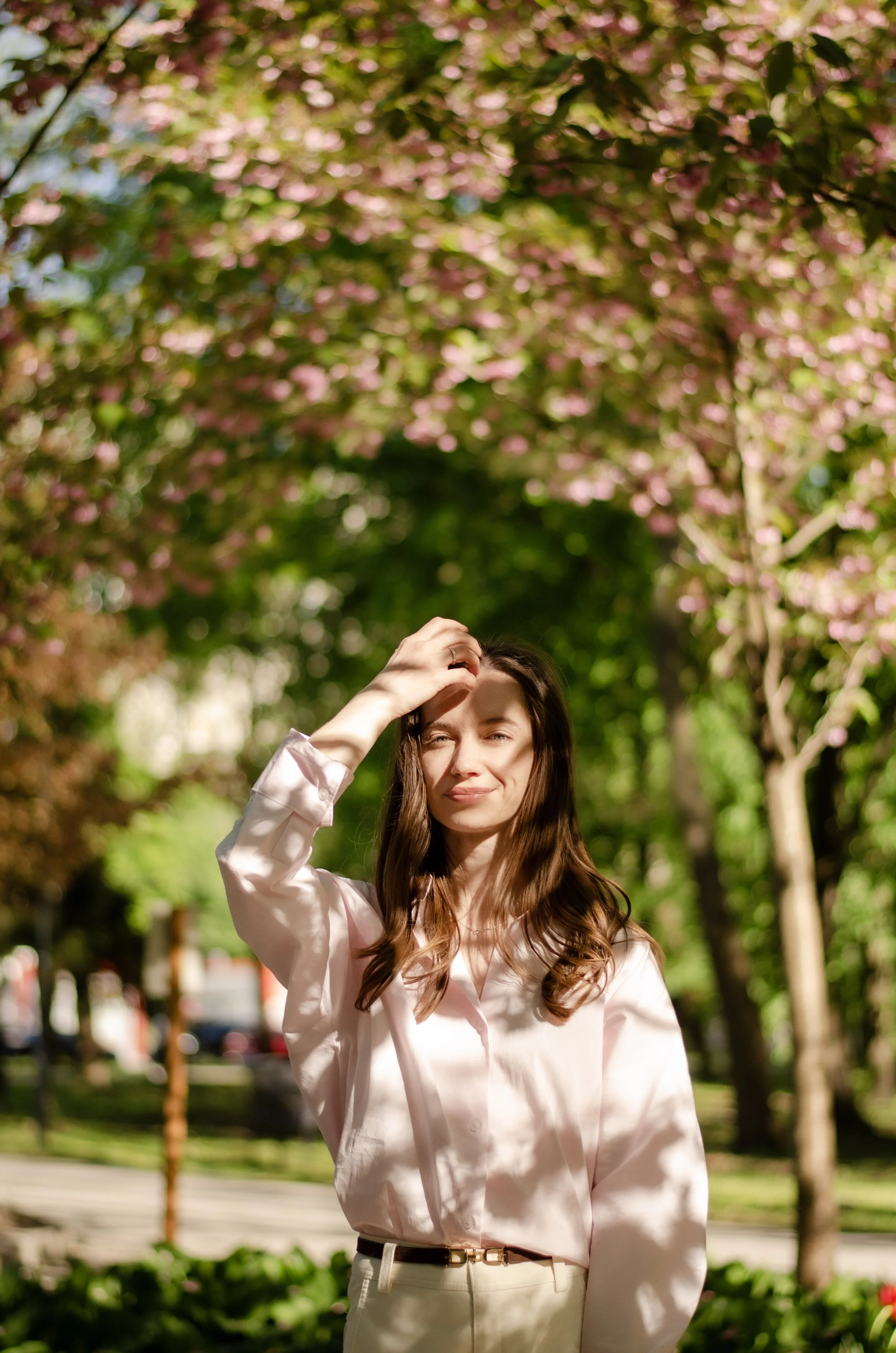 A young woman with long brown hair smiling outdoors, wearing a light pink blouse, standing in front of pink flowering trees.