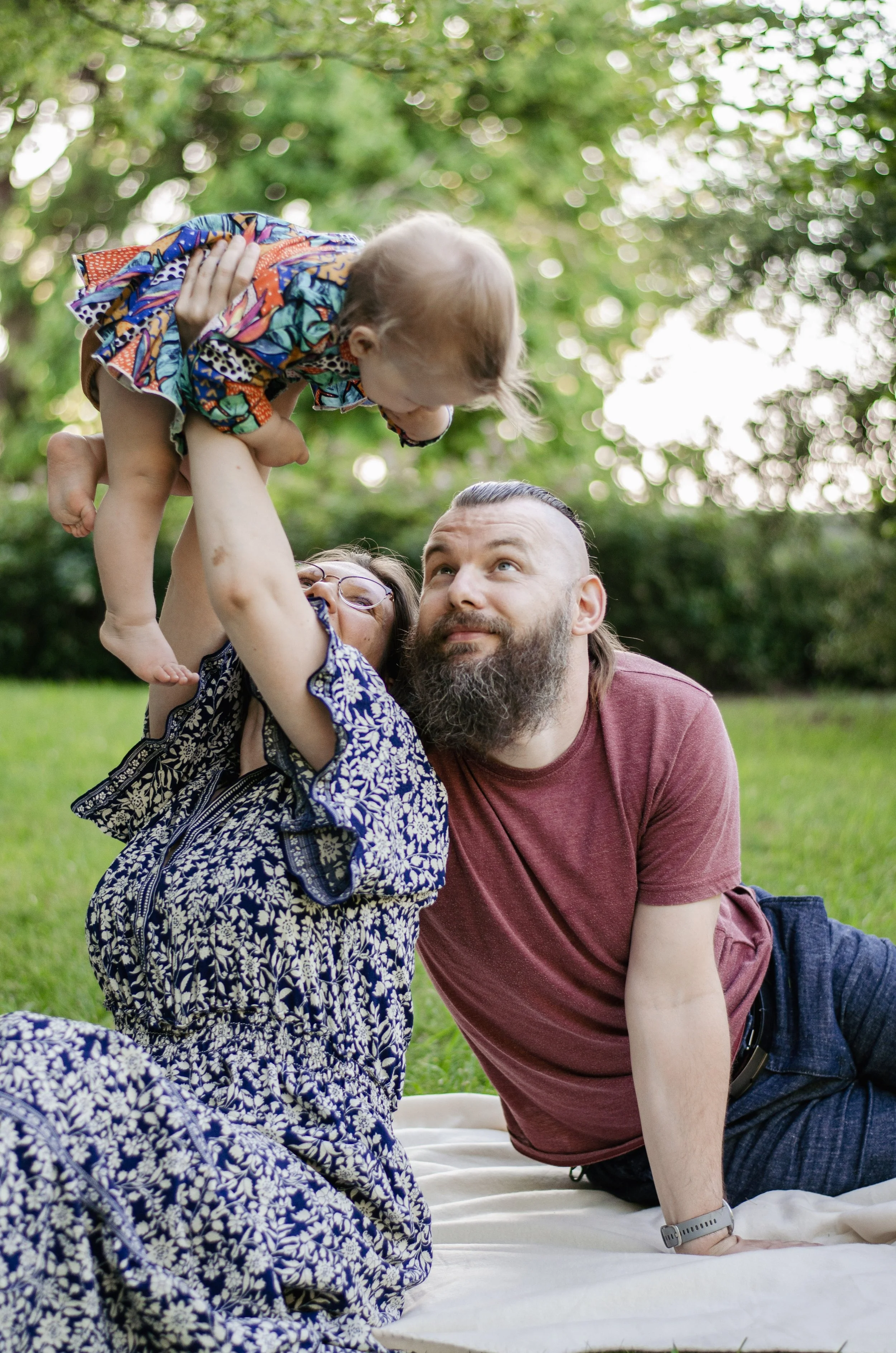 A young girl with glasses sitting on a white blanket, lifting a man, likely her father, into the air outdoors surrounded by green trees and grass. The man has long hair, a beard, and is wearing a maroon t-shirt with a fitness tracker on his wrist. The girl is wearing a blue and white patterned dress.