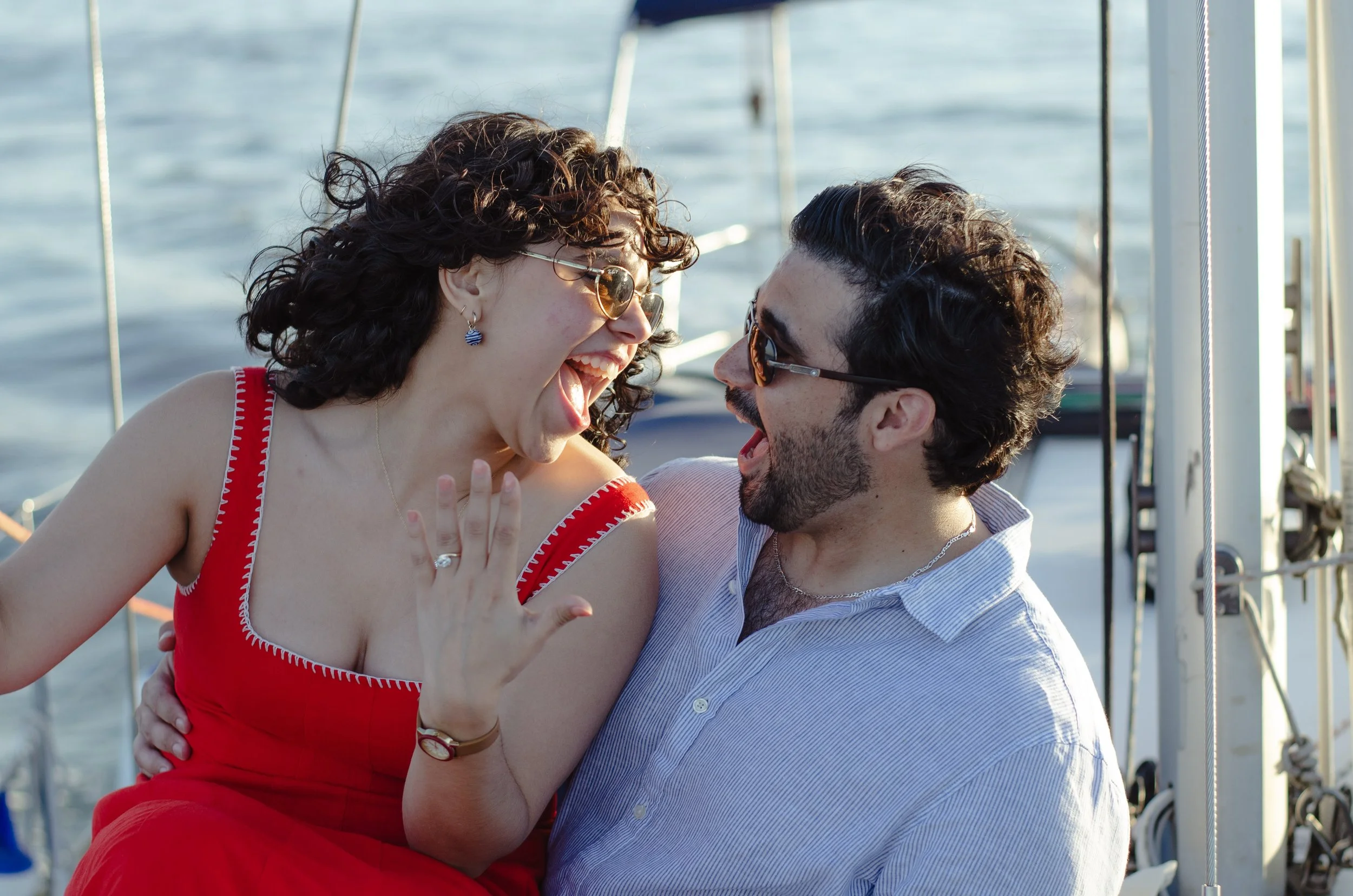 A joyful couple is hugging and laughing on a boat with water and parts of the boat's railing visible in the background.