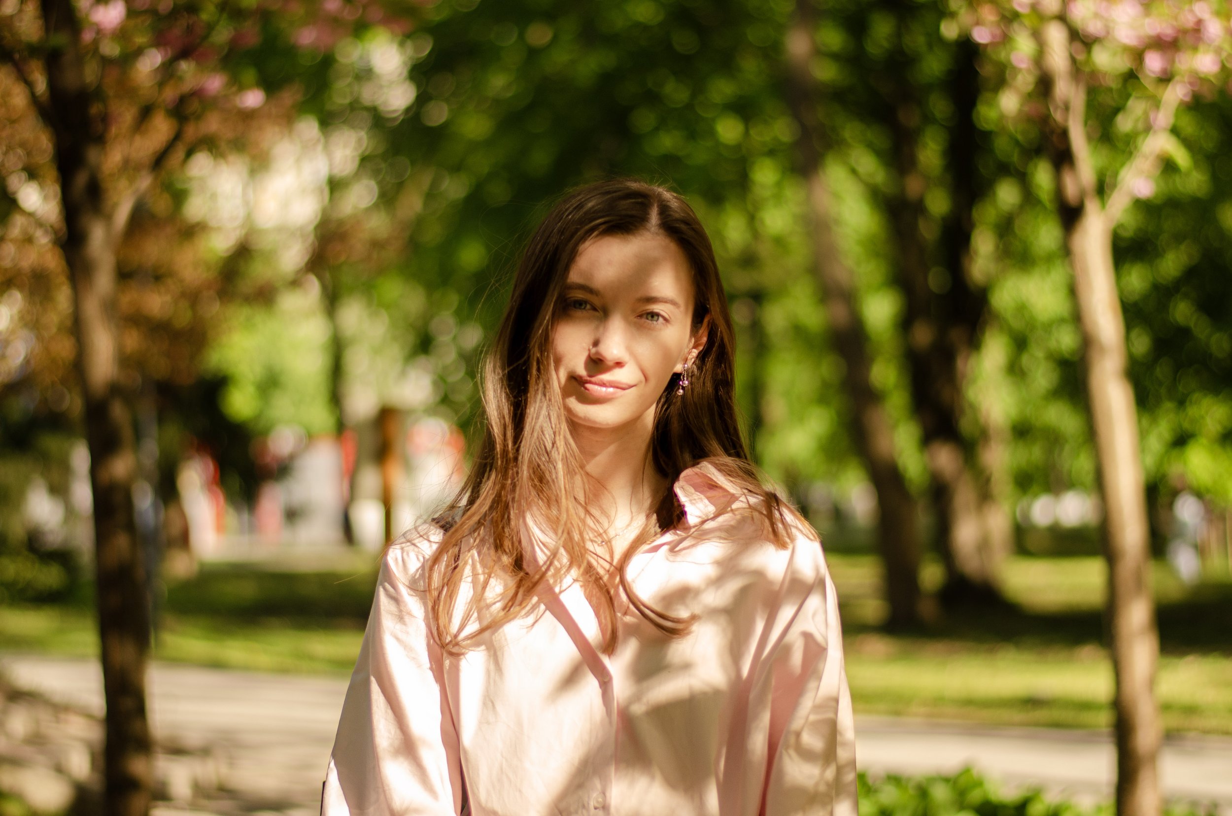 A young woman with long brown hair, wearing a pink blouse and earrings, outdoors in a park with trees and sunlight filtering through the leaves.