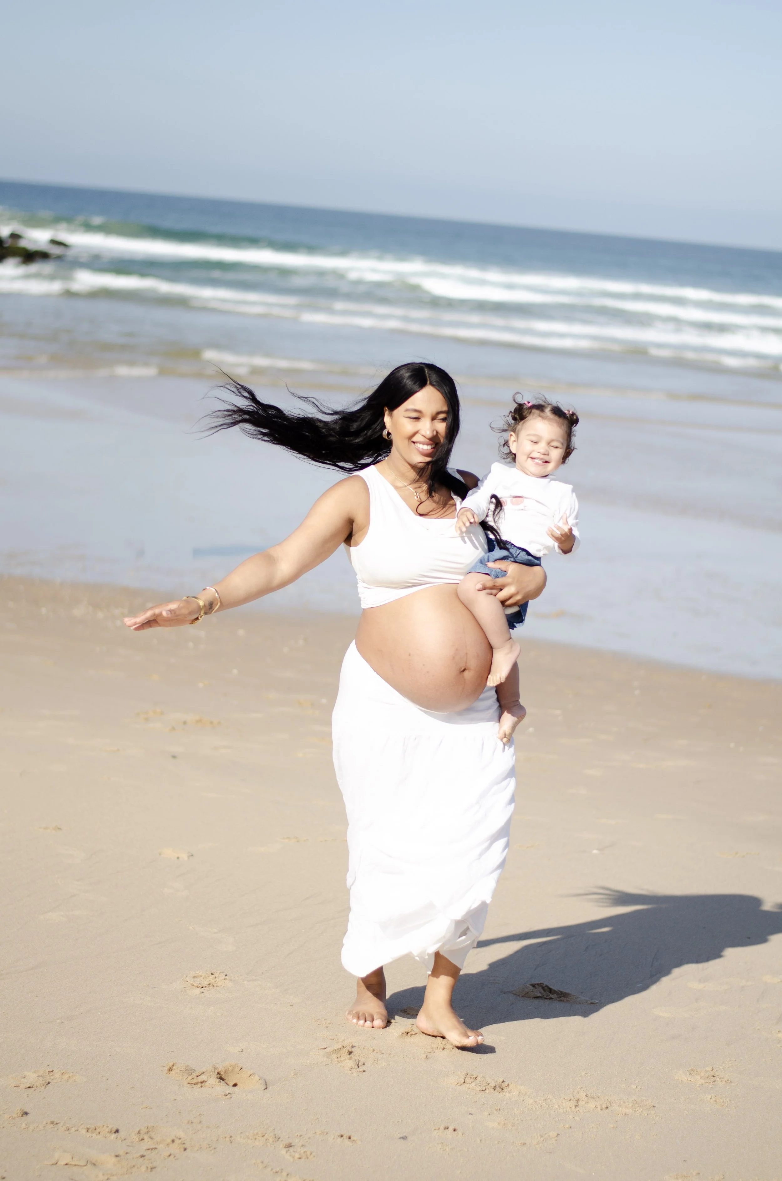 A pregnant woman smiling and holding a young girl on a sandy beach with ocean waves in the background.