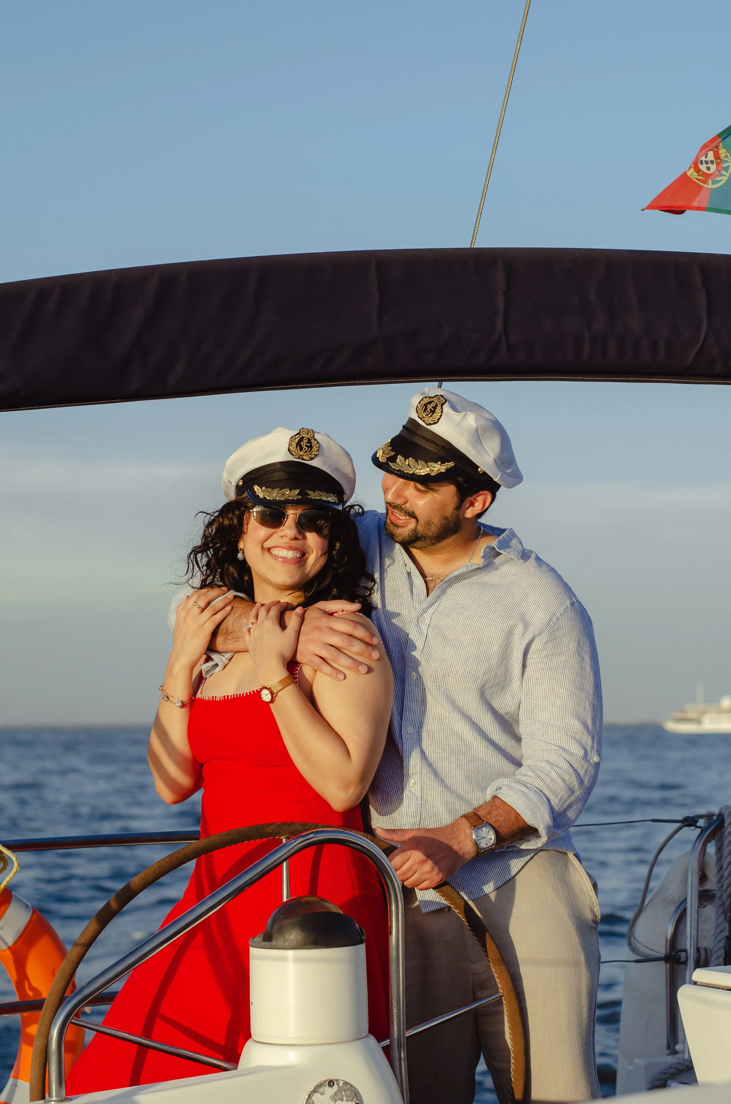 A man and woman on a boat, wearing captain's hats, smiling and embracing each other, with the ocean in the background and a ship in the distant horizon.