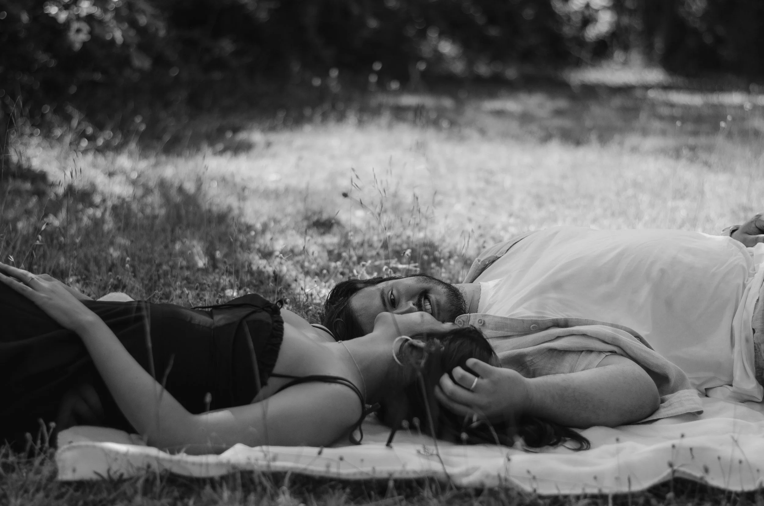 Two people lying on the grass outdoors, smiling and looking at each other. One person is wearing a white shirt and the other a dark shirt. The background consists of trees and sunlight filtering through the leaves. The image is in black and white.